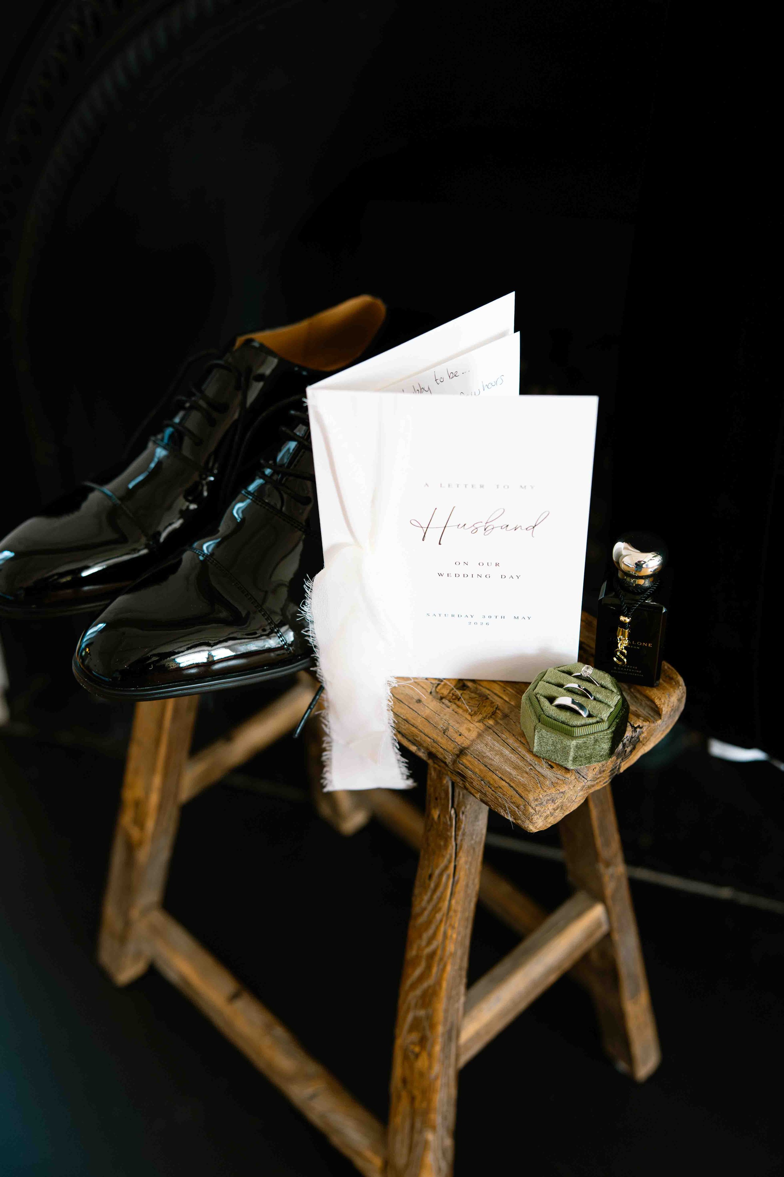Black shiny shoes, wedding invitation on a rustic wooden stool, small perfume bottle, and small box with rings, dark background.