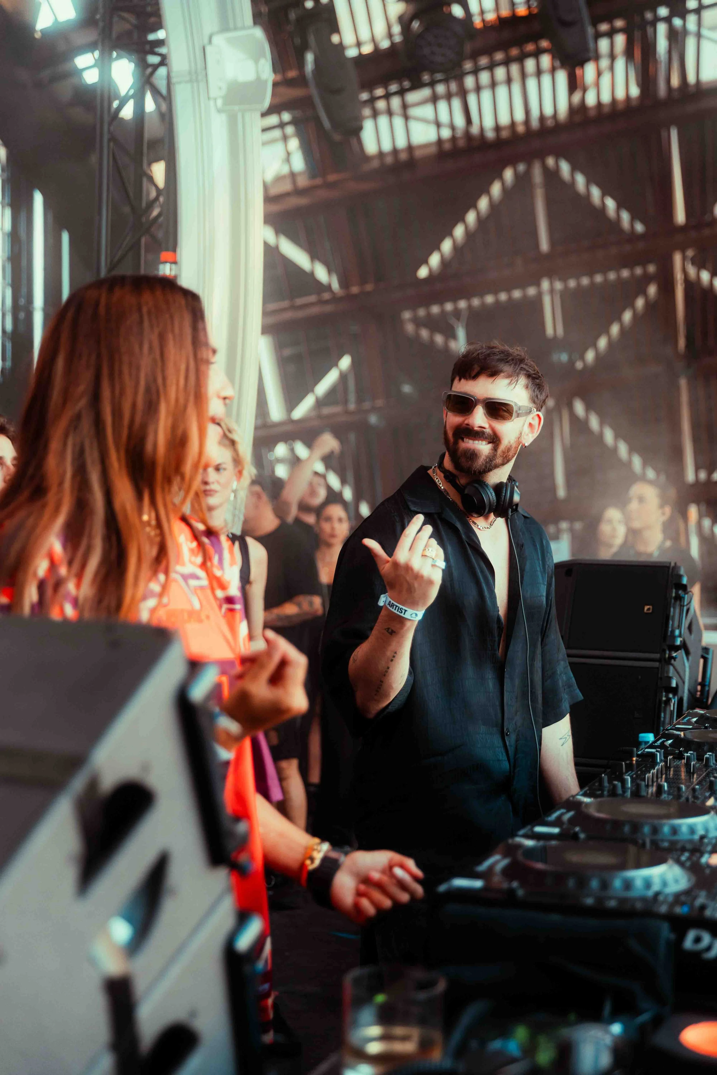 A DJ with glasses and headphones around his neck smiling at a woman at a music event inside an industrial-style venue with exposed beams.