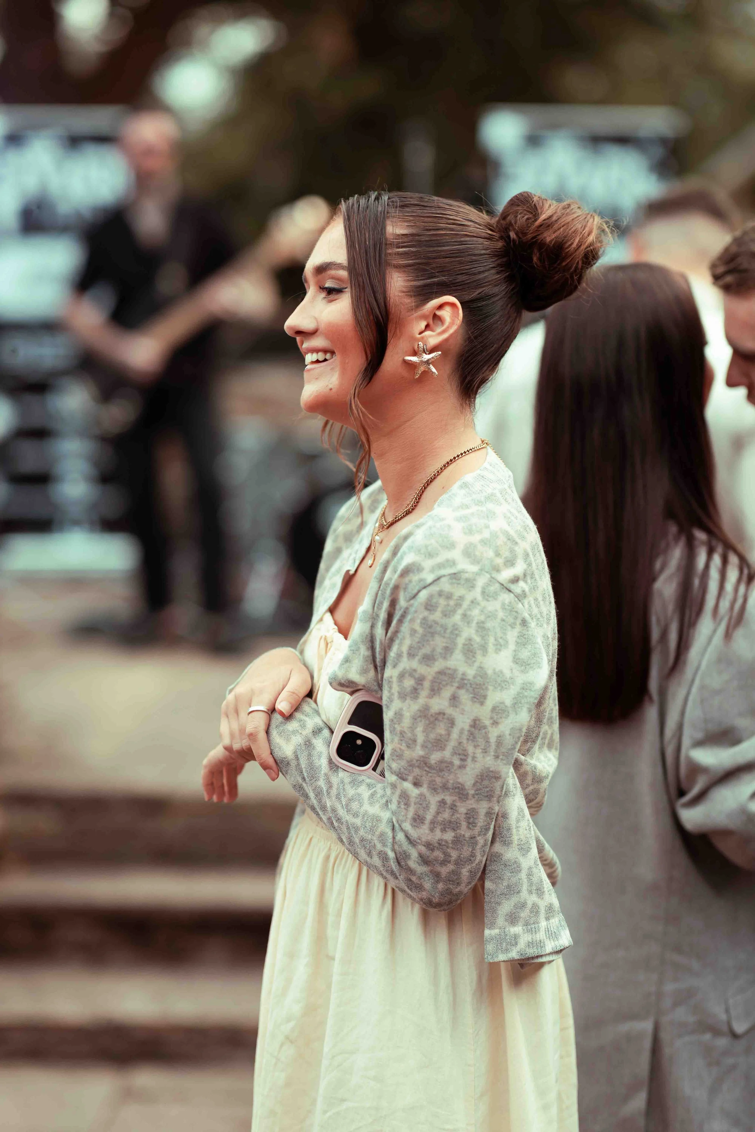 A woman stands outdoors with her arms crossed, smiling and wearing starfish-shaped earrings, a light-colored patterned jacket, and a yellow dress, with a smartphone tucked into her sleeve. In the background, slightly out of focus, there is a man and 