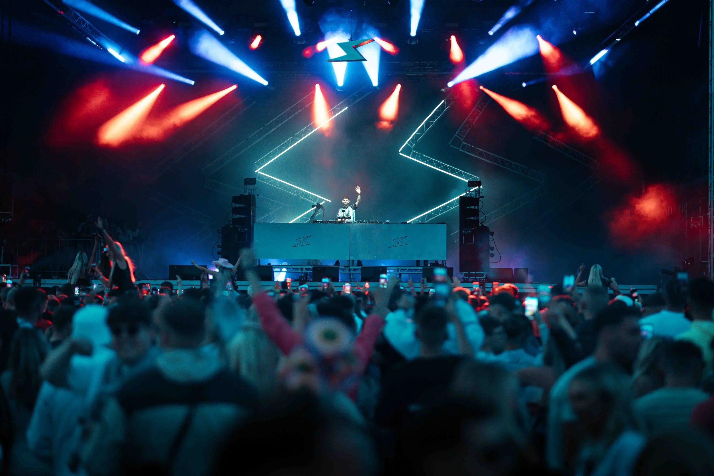 A DJ performs on stage with colorful lights and a crowd watching at a concert.