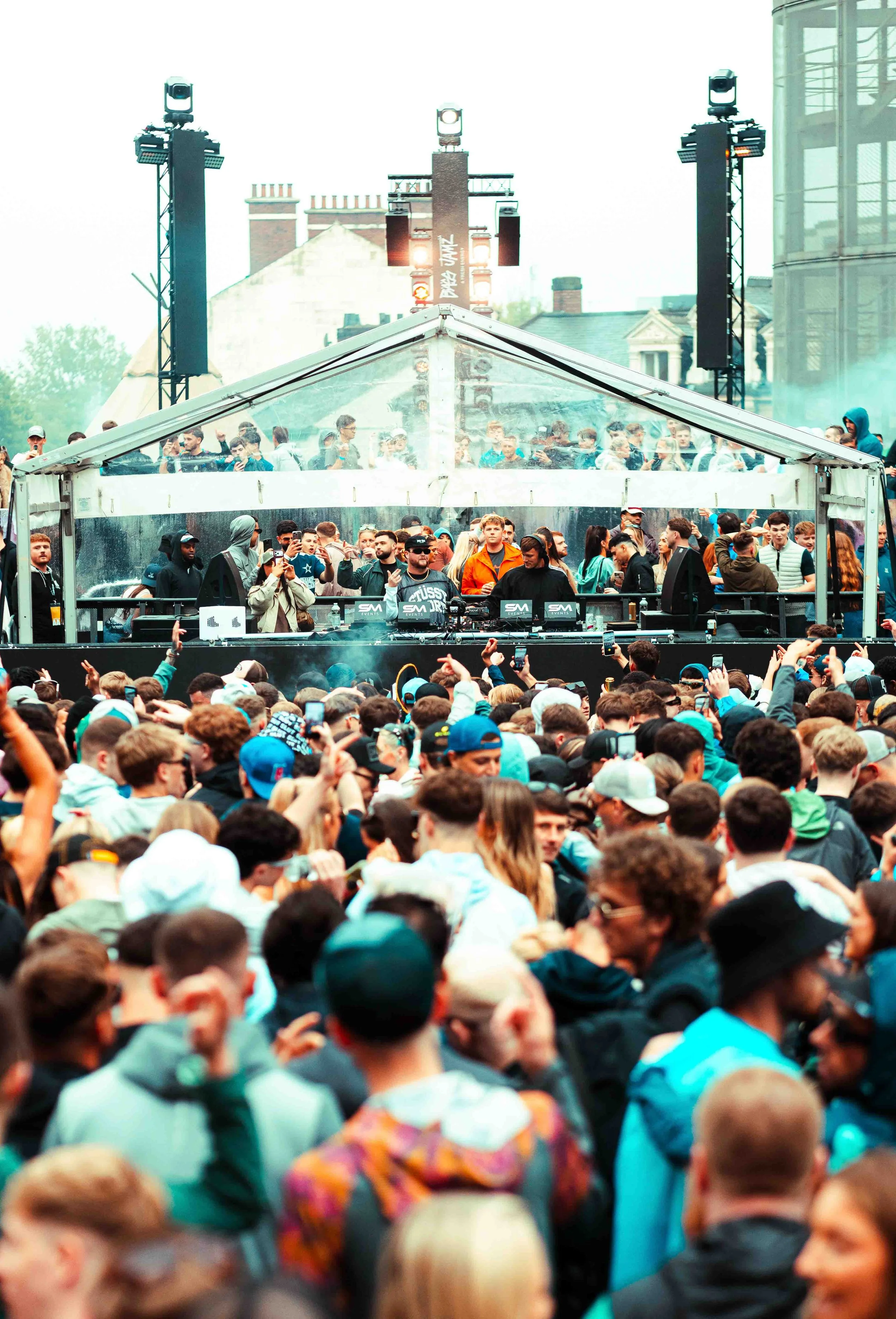 Crowd of people at an outdoor music event with a stage, DJ, and DJ equipment.