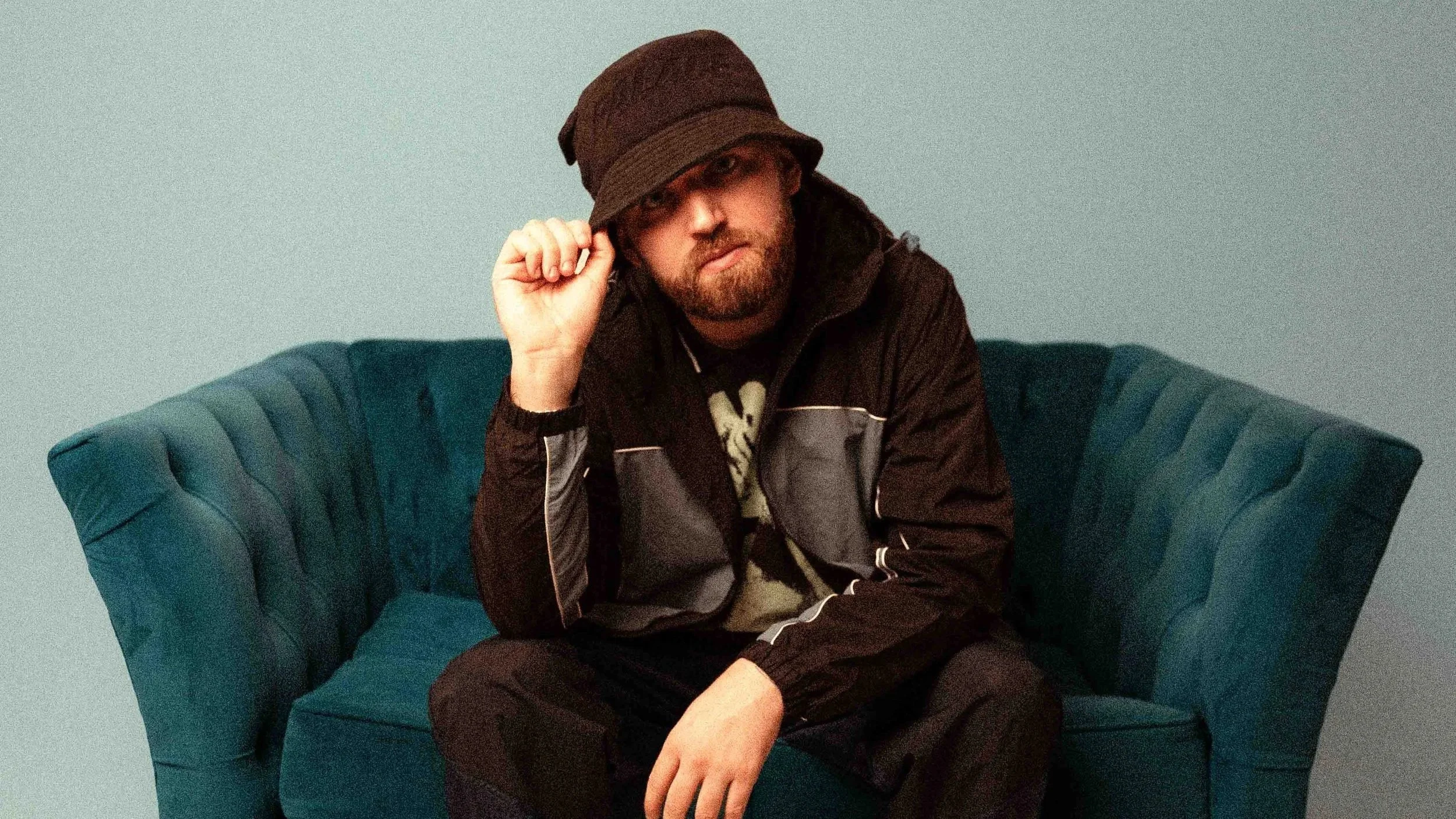 A man with a beard wearing a brown bucket hat and black jacket sitting on a teal upholstered armchair against a plain wall.