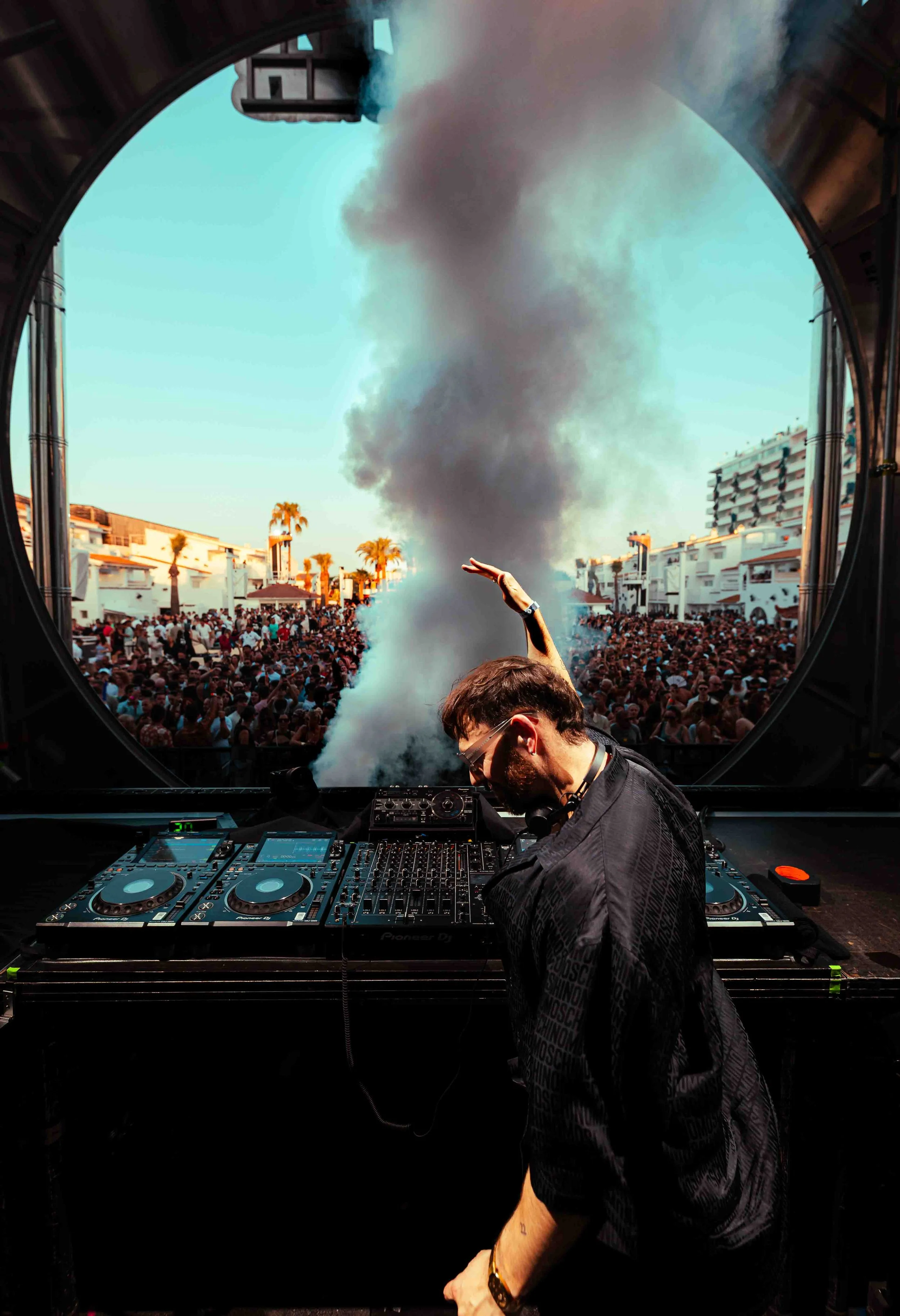 A DJ wearing glasses and a black shirt, mixing music on a professional DJ set while a large crowd dances outdoors. Smoke rises in the background with buildings and palm trees visible under a clear sky.