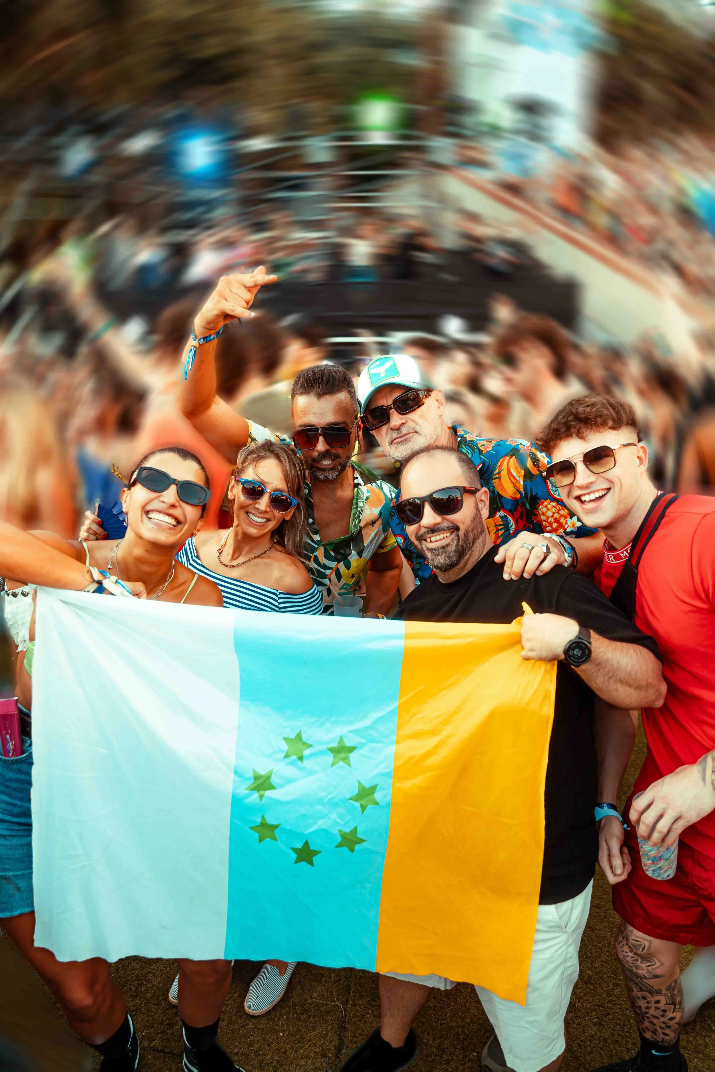 A group of smiling people holding a flag with green stars, celebrating at an outdoor event with a crowd and stage in the background.