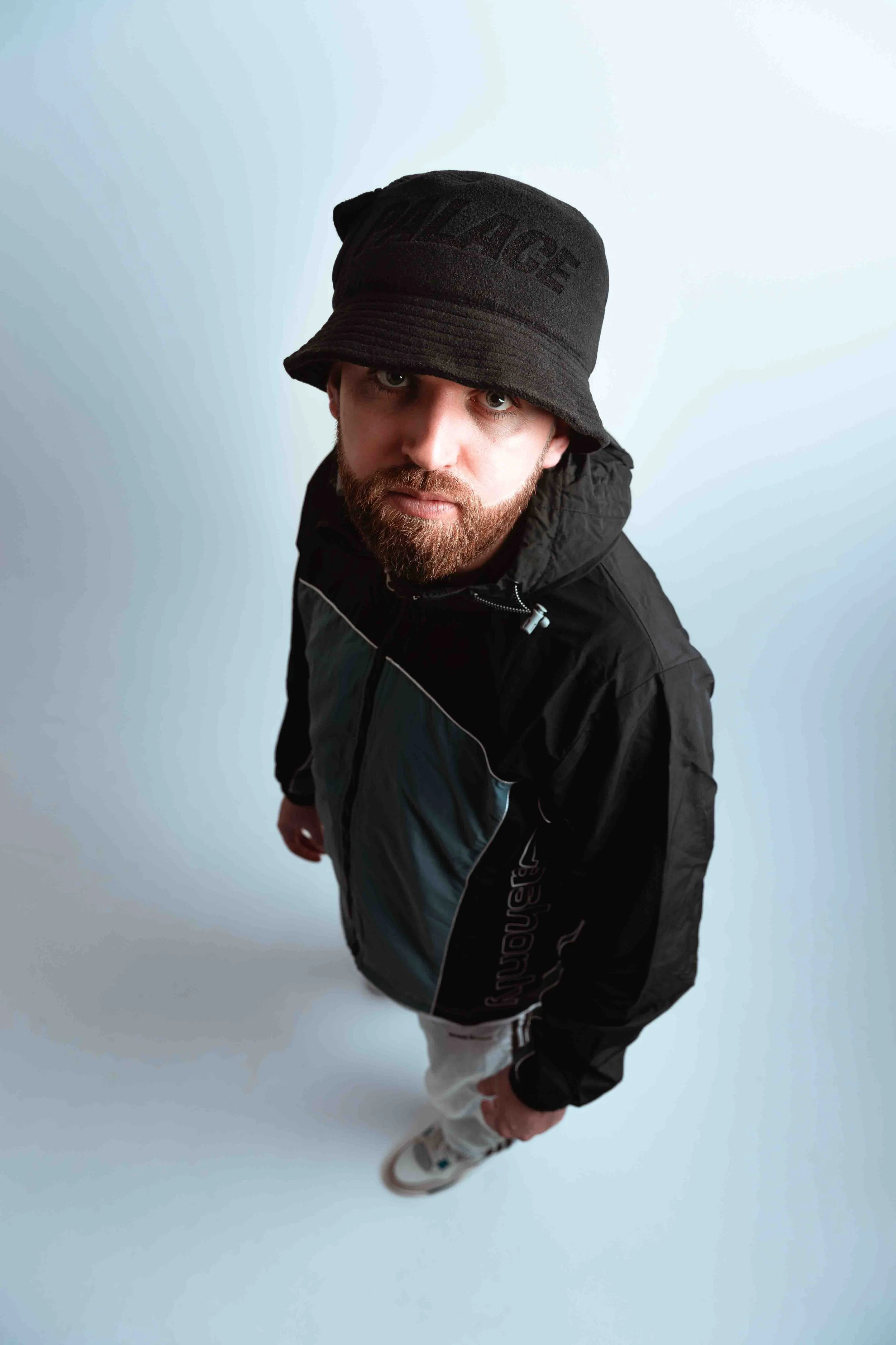 Gaskin with a beard wearing a black bucket hat, black jacket, and gray pants, looking directly at the camera, standing on a light-colored floor.