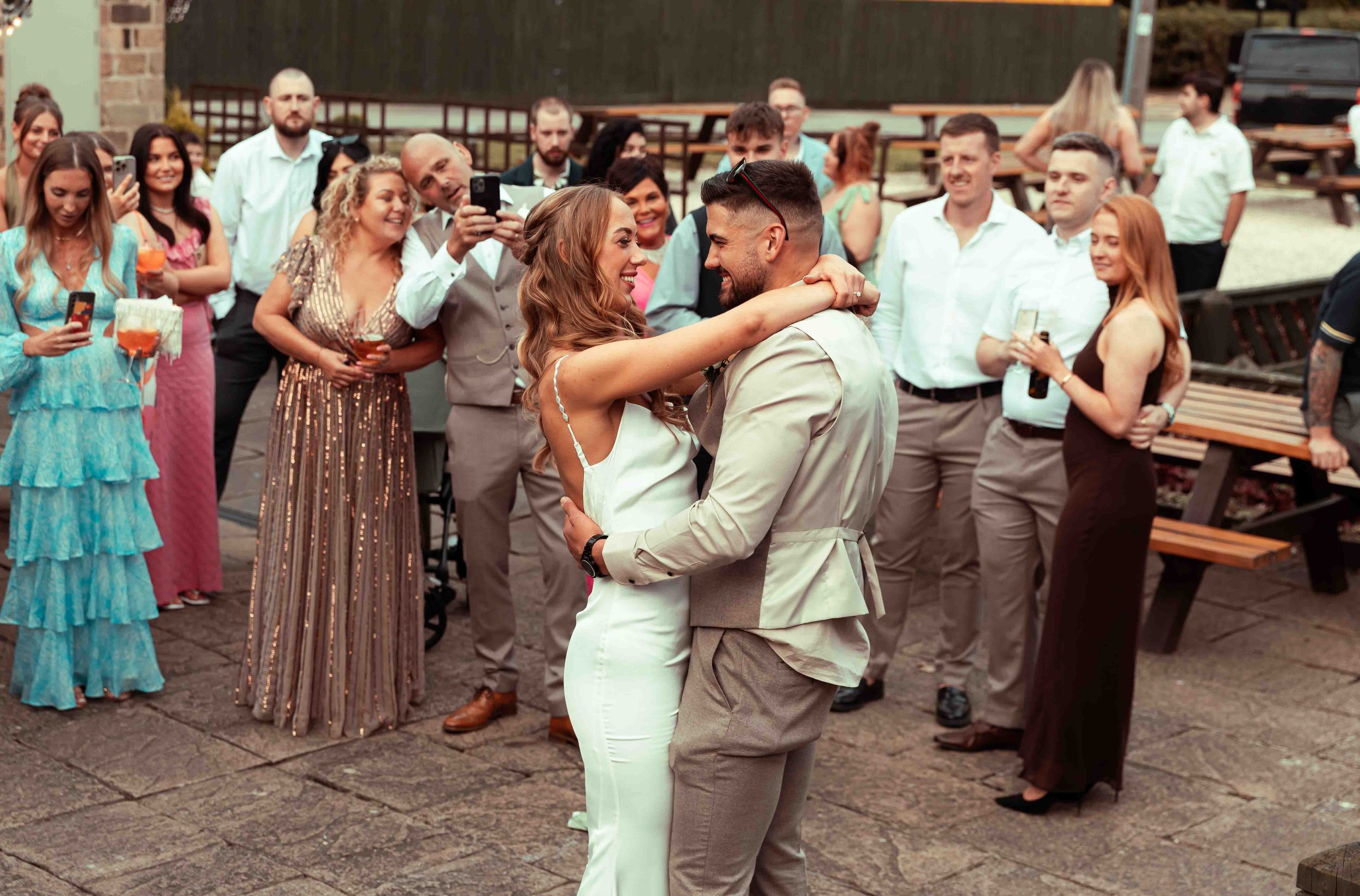 A couple is dancing closely together while surrounded by friends at an outdoor gathering or party. The woman has long, wavy hair and is wearing a white outfit, and the man has short dark hair, a beard, and is wearing a light-colored suit. They are sm