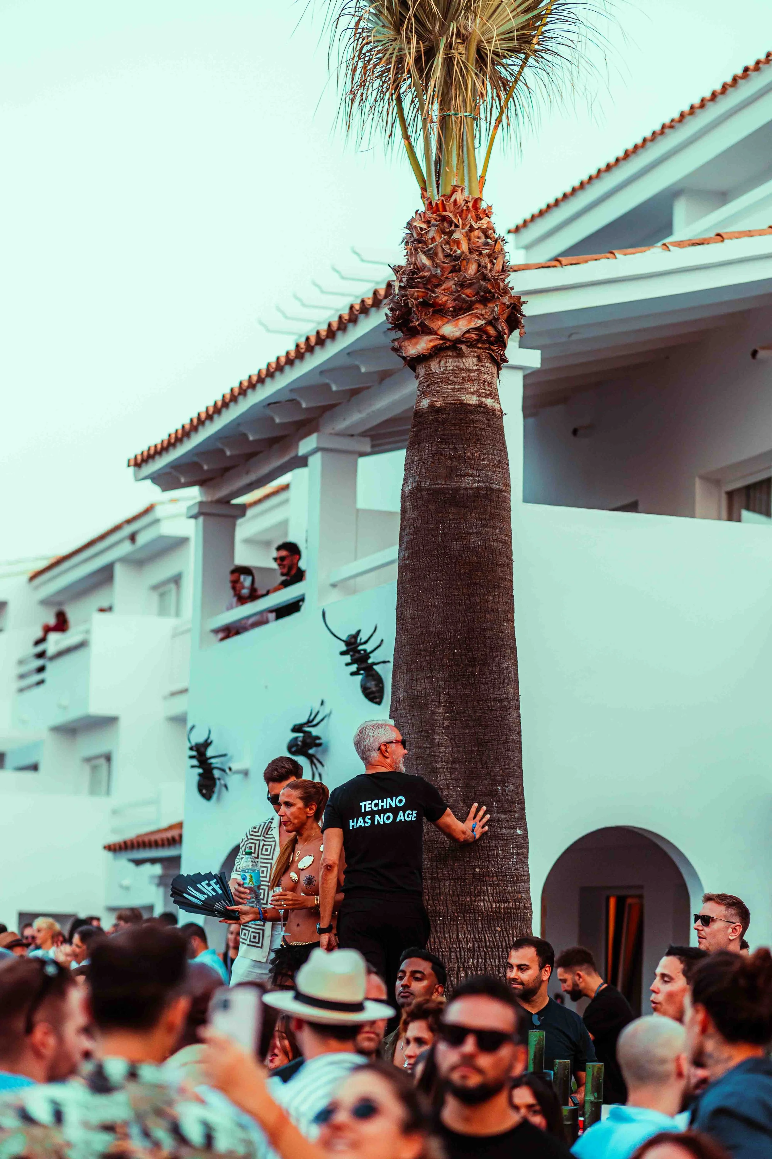 A crowded outdoor scene with many people gathering under a large palm tree in front of a white building with orange-tiled roof. Some individuals are standing, talking, or seated, and there are decorations on the wall resembling bees or wasps.
