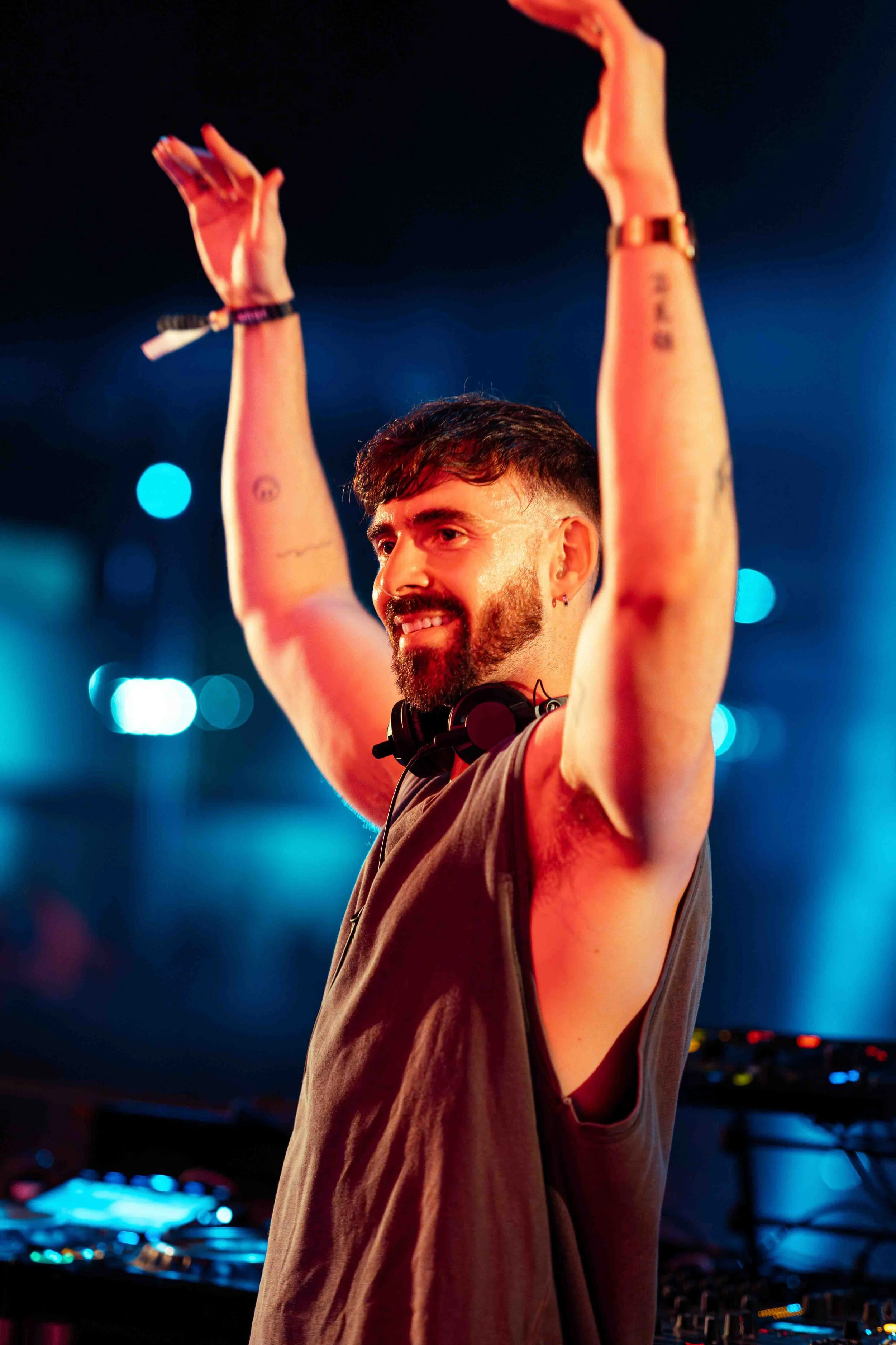 A male DJ with a beard and tattoos on his arms raising his hands in the air wearing headphones around his neck, with colorful lights and DJ equipment in the background.