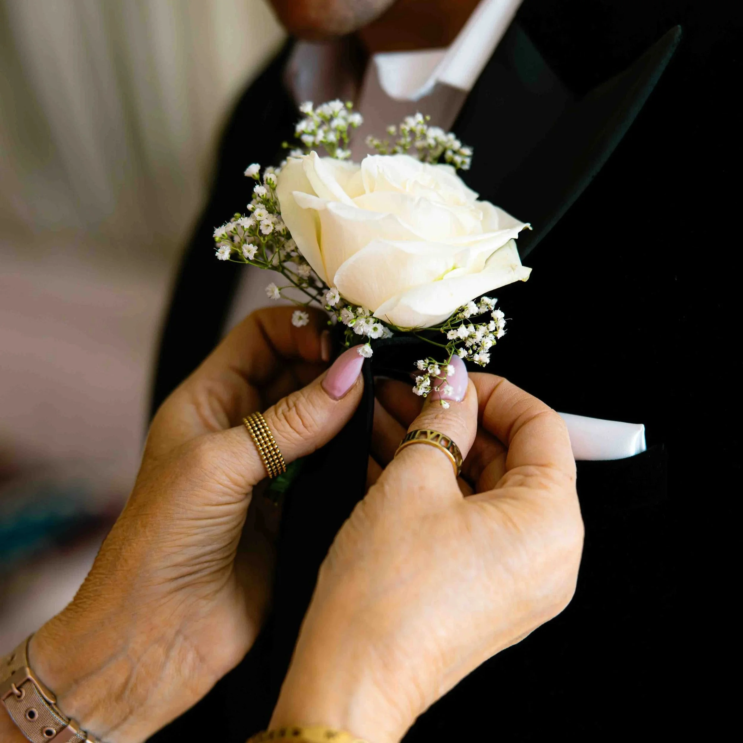 Close-up of a hand holding a bouquet with a white rose and small white flowers, with rings on the fingers and a beige watch on the wrist.