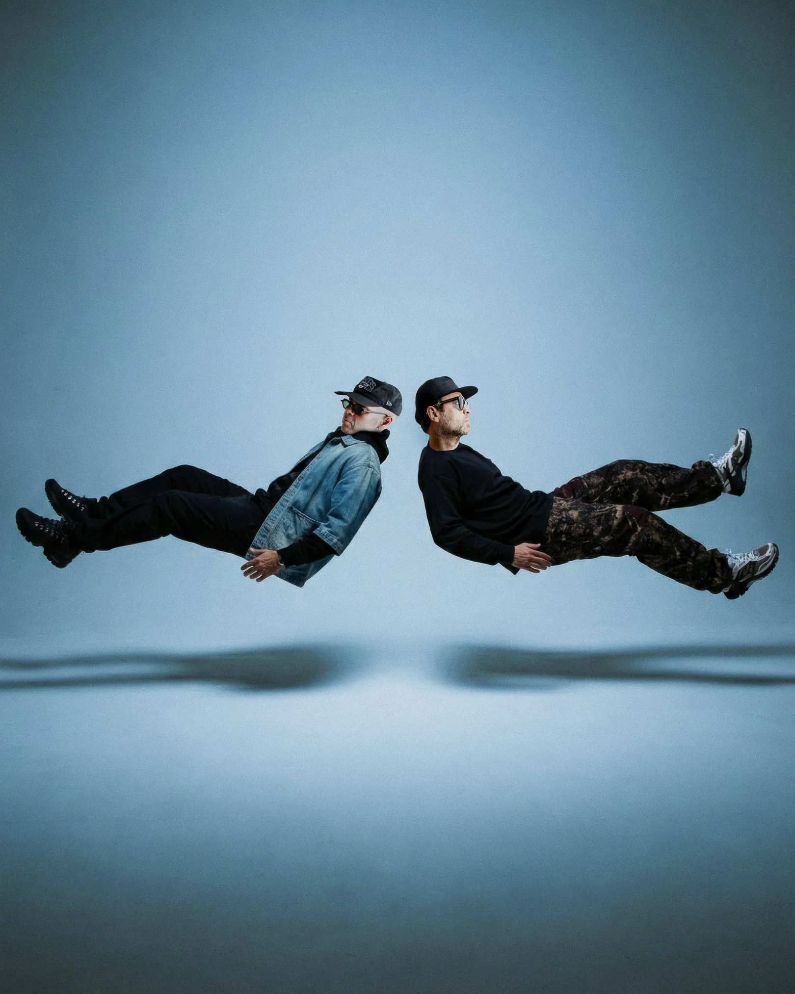 Two men sitting back to back with legs extended and lifted slightly off the ground, positioned horizontally across the image, against a plain blue background.