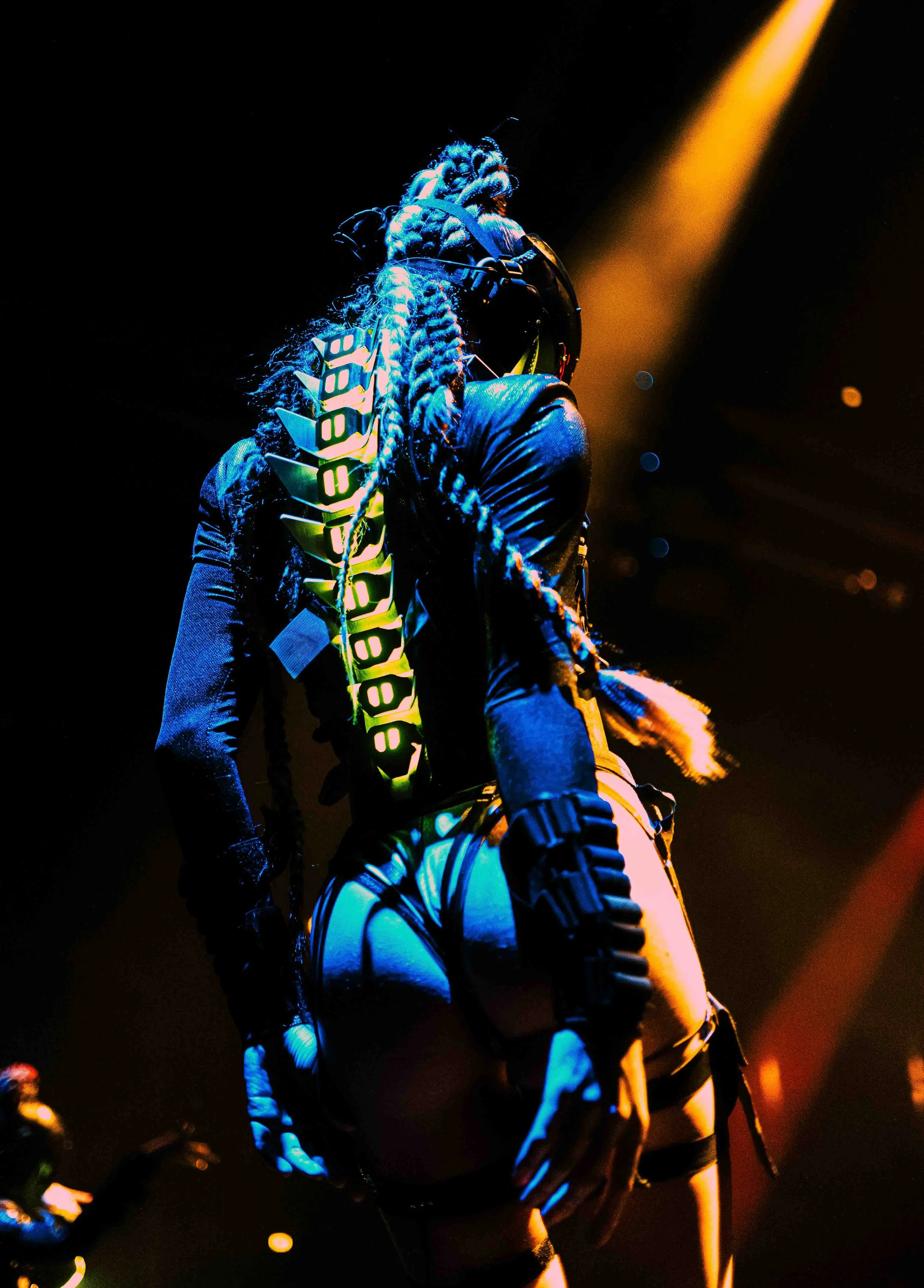 A person with long, braided hair, wearing a dark jacket and pants, performing on stage under bright, colorful lights, with large, gear-like accessories on their back and arm, in a dark setting.