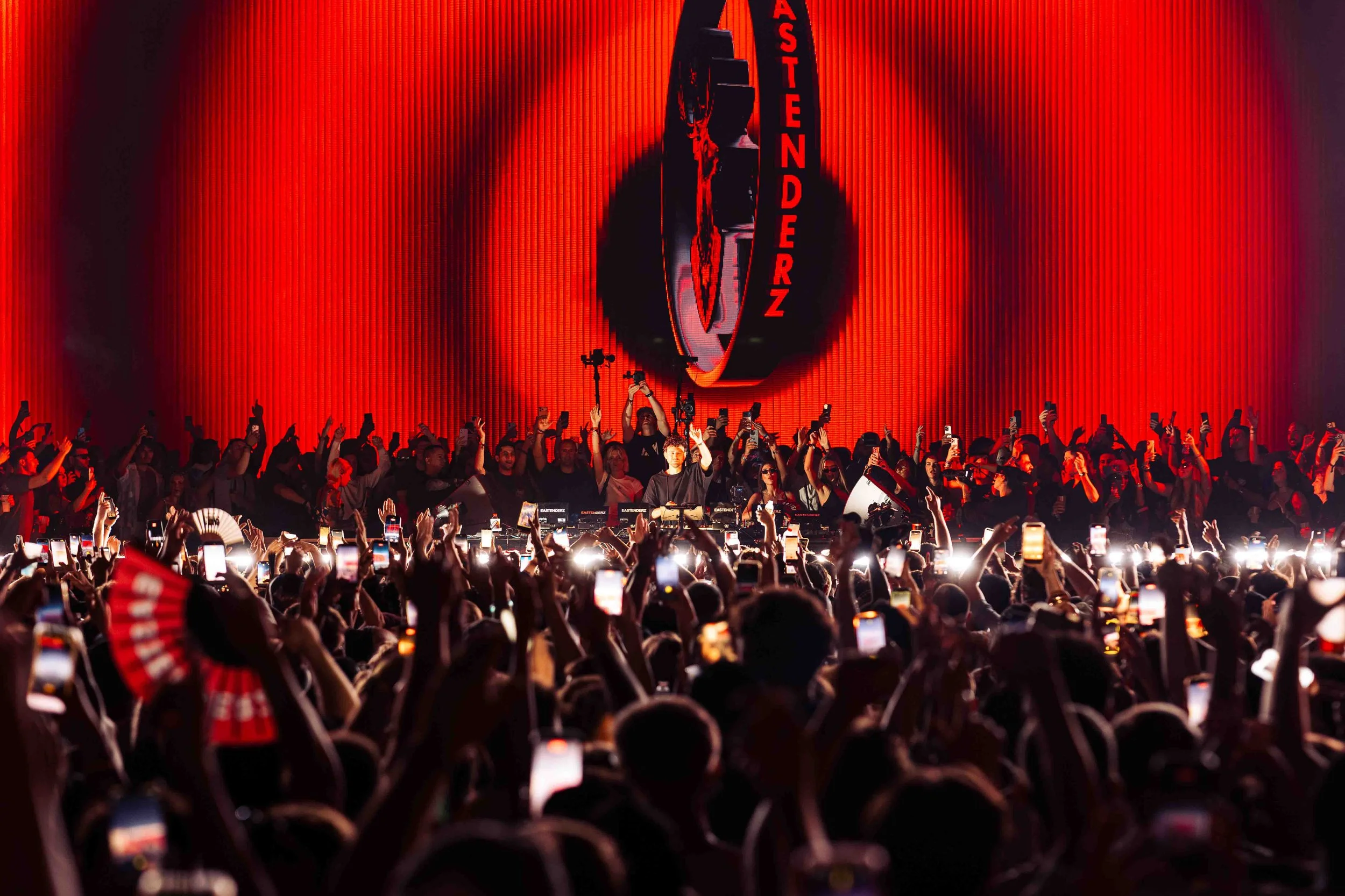 A large crowd of people at a concert or event with many holding up their phones. There is a stage in the background with a red-lit sign that reads 'EASTENDERZ'. The stage is framed by a large circular structure and red curtains.
