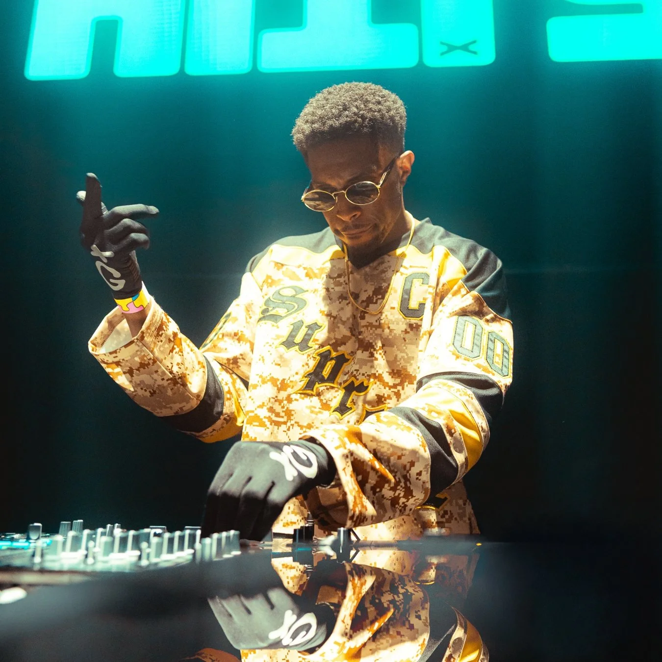 A DJ wearing camouflage-patterned jacket with various patches and black gloves is performing on stage with turntables, illuminated by green lights. The DJ is concentrating, wearing sunglasses and a gold chain.