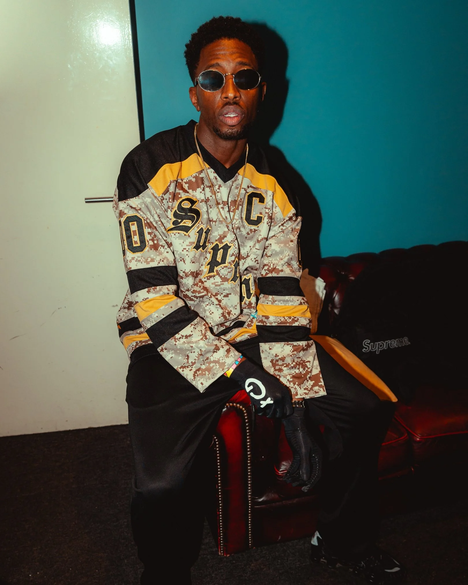 A man wearing round sunglasses, a camouflage sweatshirt with yellow and black accents, and black pants, sitting on a chair against a teal wall. He has a chain necklace and several colorful bracelets. There's a black Supreme bag and a black jacket with a red interior nearby.