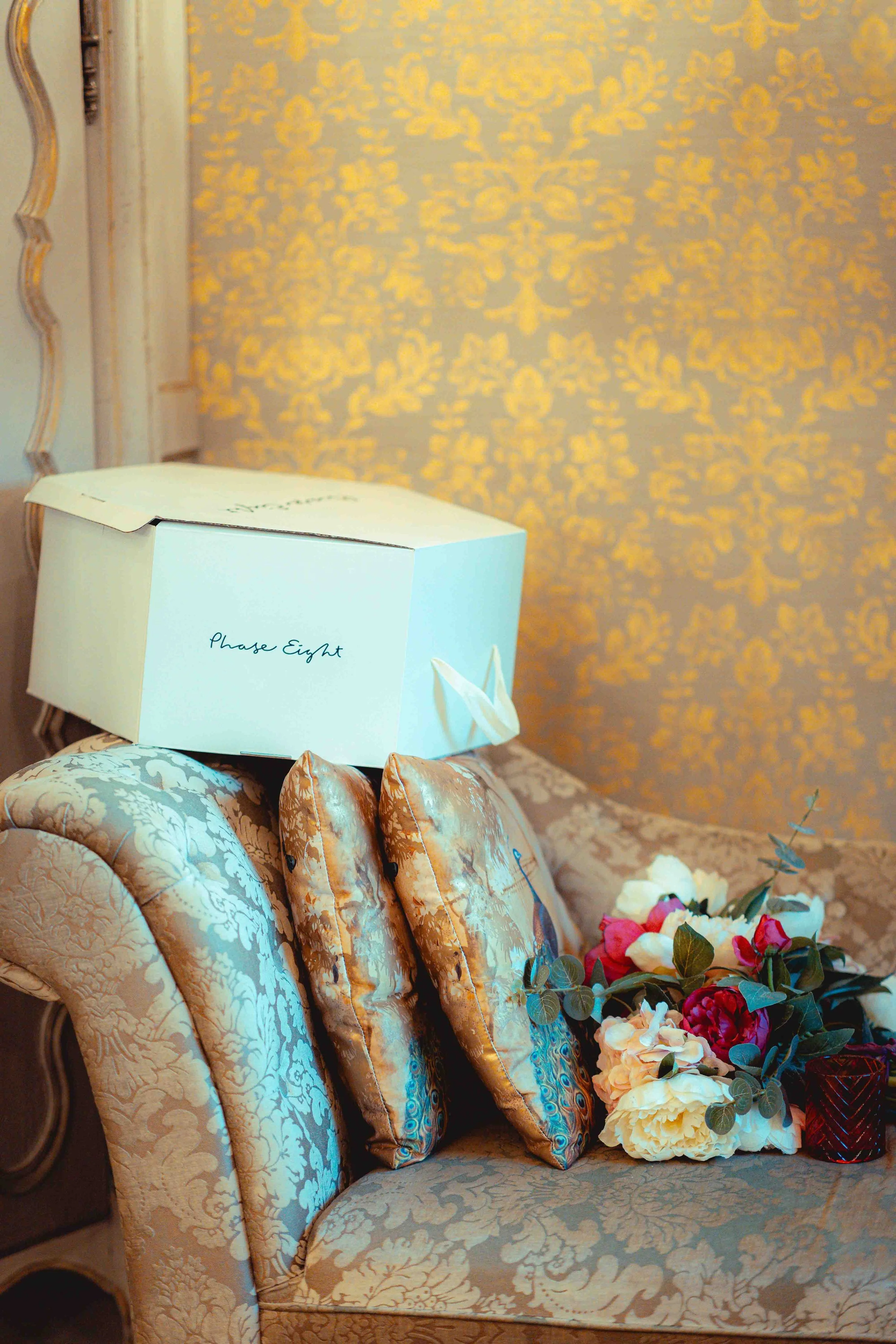 A vintage-style cream-colored loveseat with floral pattern upholstery, placed against a gold damask-patterned wall. On the seat, there is a white box with the words "Phase Eight" printed on it and a bouquet of pink, white, and yellow flowers.