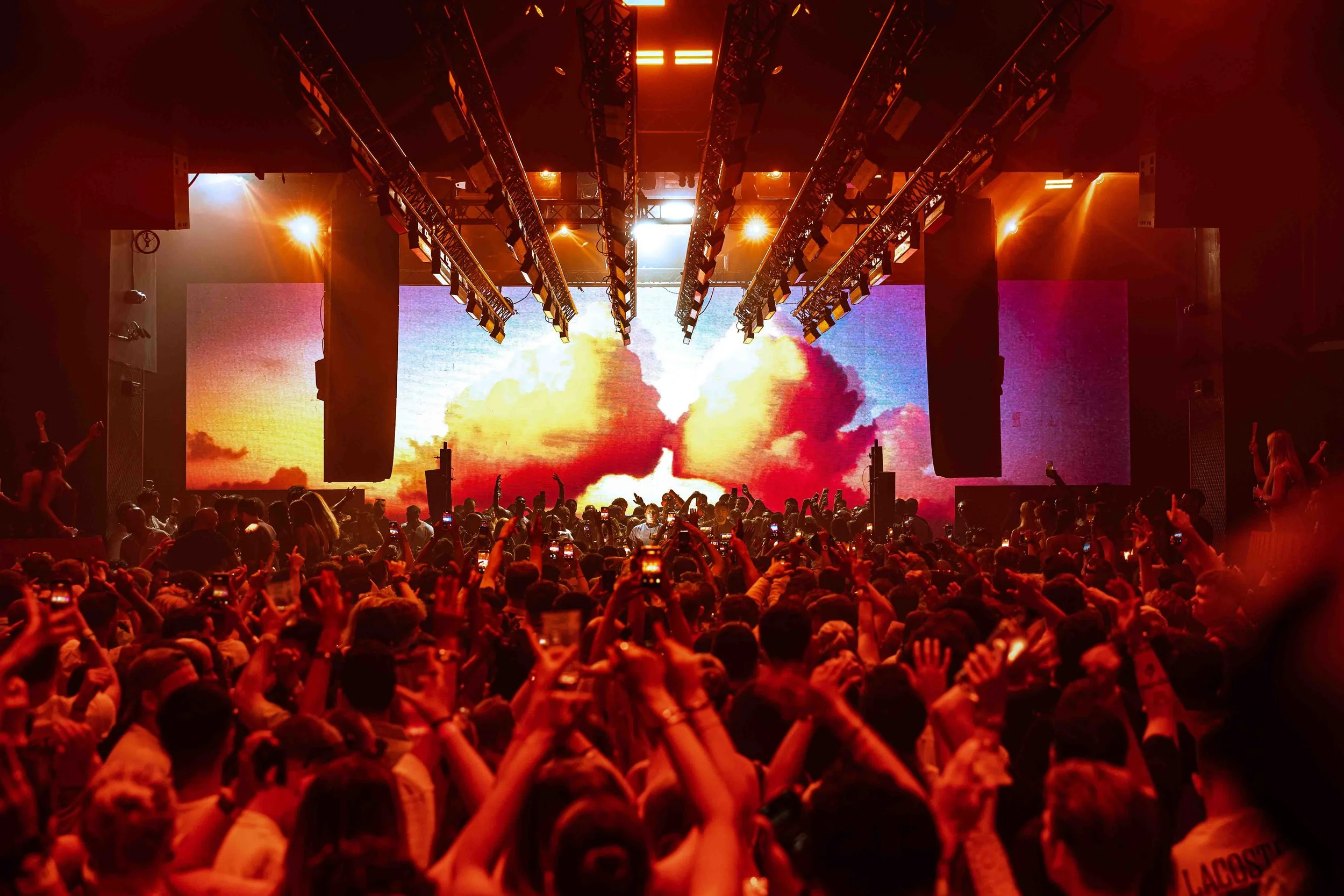Crowd of people at a concert or event with stage lights and large digital screens displaying a sunset or sunrise background.