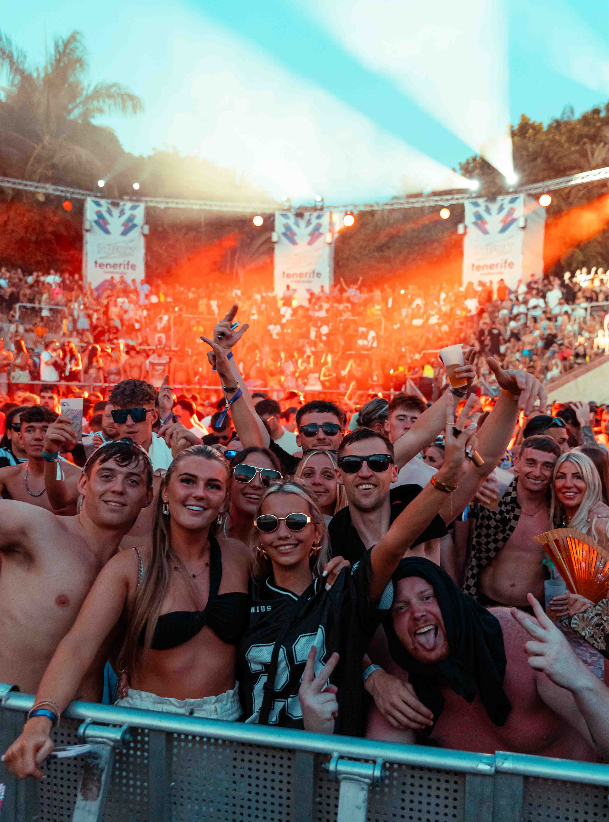 Crowd of young people at an outdoor concert or festival during sunset, with stages and banners in the background that say "tenerife" and with palm trees visible, some people raising their hands and smiling.