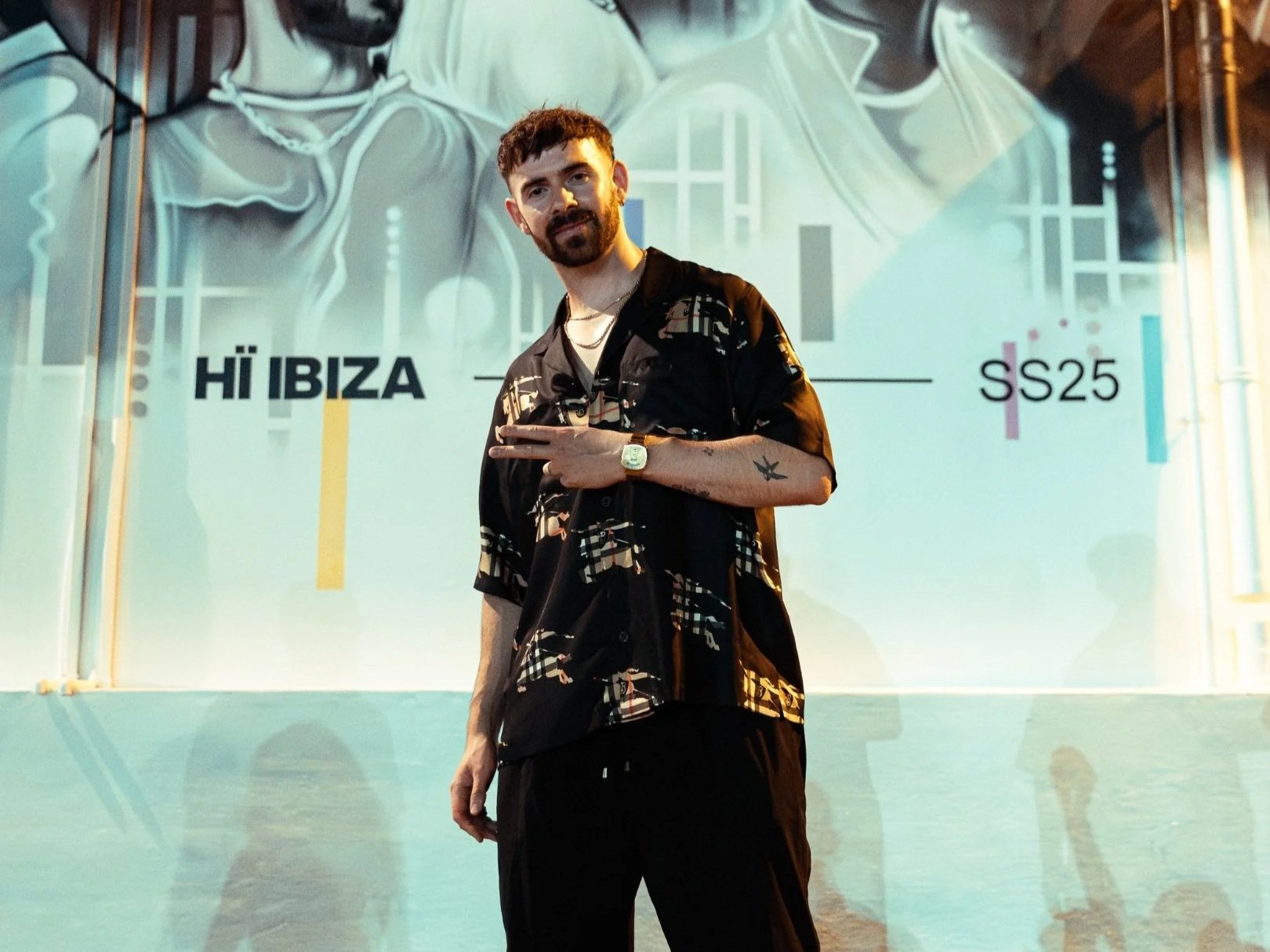 A man with dark hair and beard posing in front of a colorful backdrop with the words 'HI IBIZA' and 'SS25'. He is wearing a black patterned shirt, black pants, a watch, and necklaces, and is making a peace sign with his right hand.