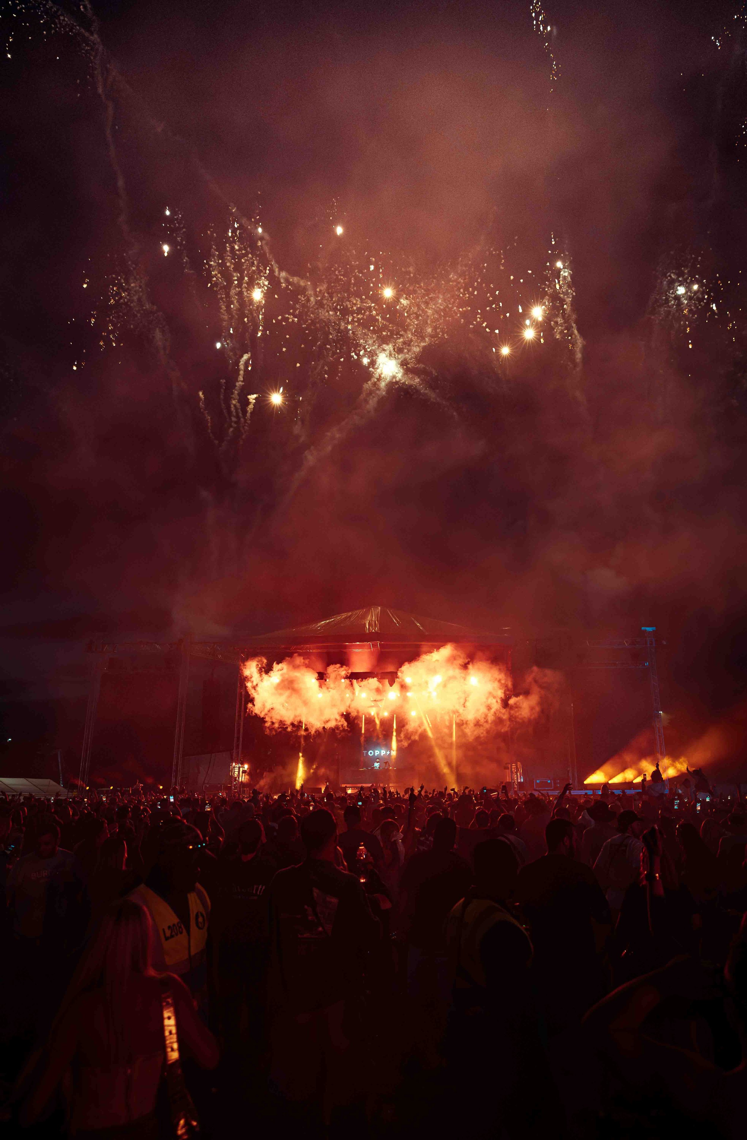 Nighttime scene at a concert or fireworks show with a large crowd, a stage with bright lights, and fireworks illuminating the sky overhead.