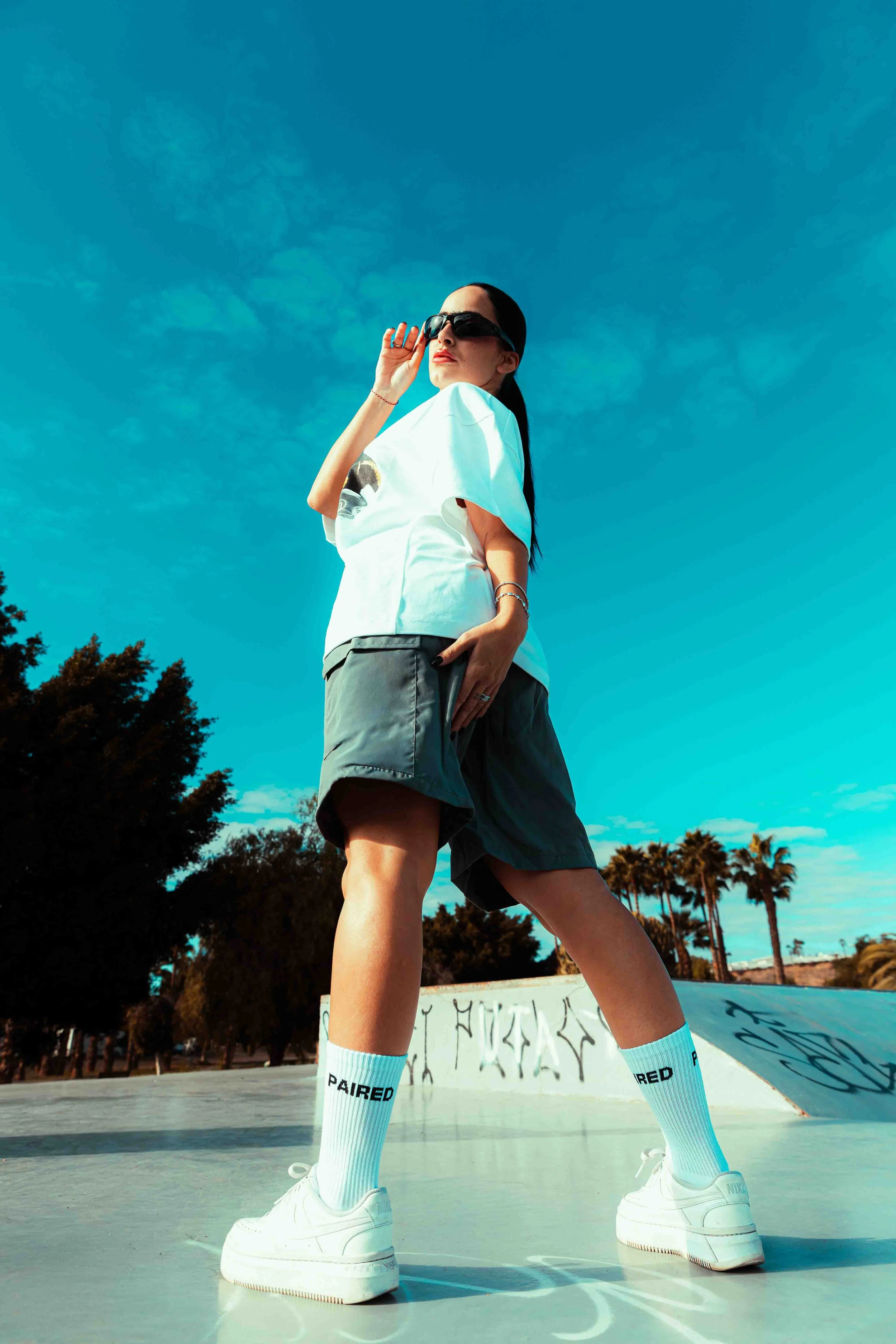 A woman wearing white Nike sneakers, white socks with black and blue text, gray shorts, a white t-shirt, and black sunglasses stands in a skate park with trees and a blue sky in the background.