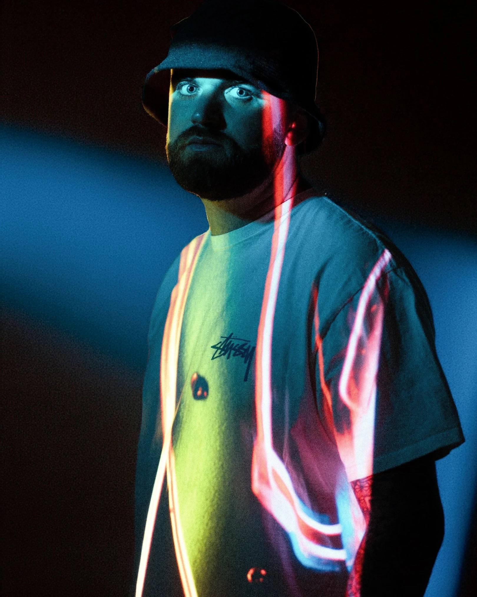 A man with a beard wearing a bucket hat and a white T-shirt with the 'Stussy' logo is illuminated by colorful neon lights in a dark setting.