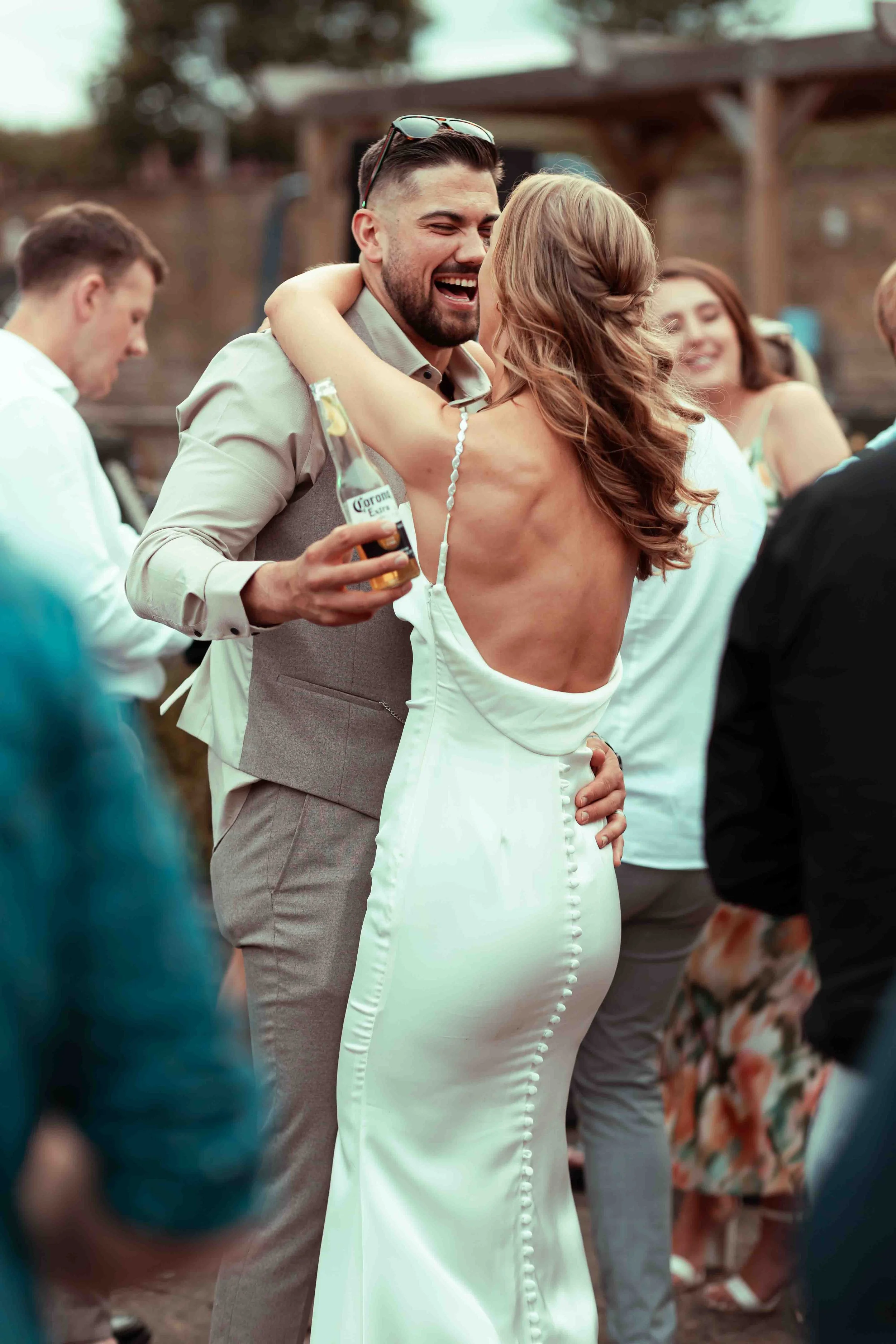 A man and woman embrace at a lively outdoor wedding reception, with the woman in a white wedding dress and the man in a suit, holding a beer, and other guests in the background.