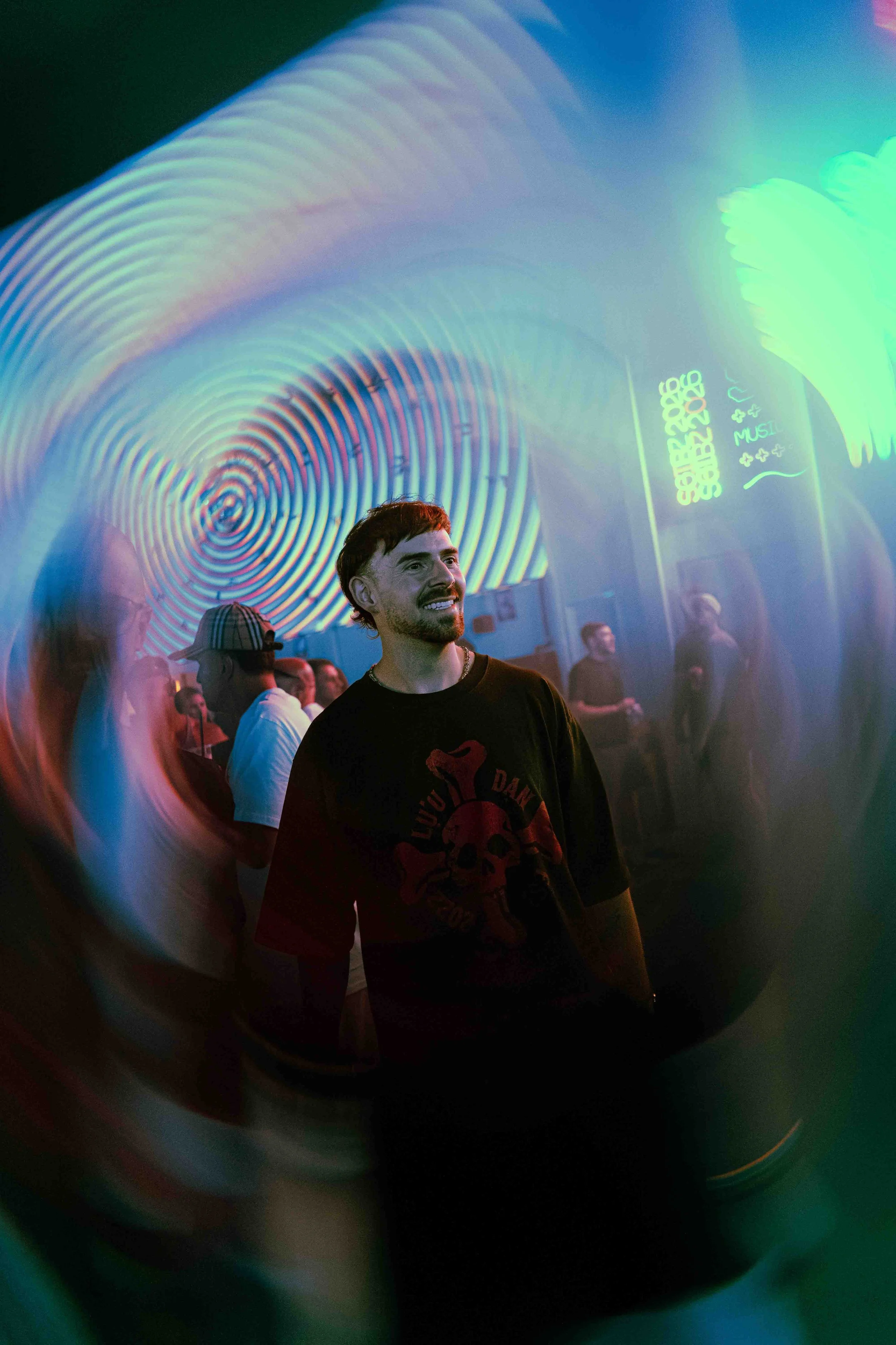 A man with dark hair and a beard is smiling and looking to his left, in a dimly lit room with multicolored lighting and multiple people in the background. The photo is taken through a circular, rainbow-colored reflective or transparent object.