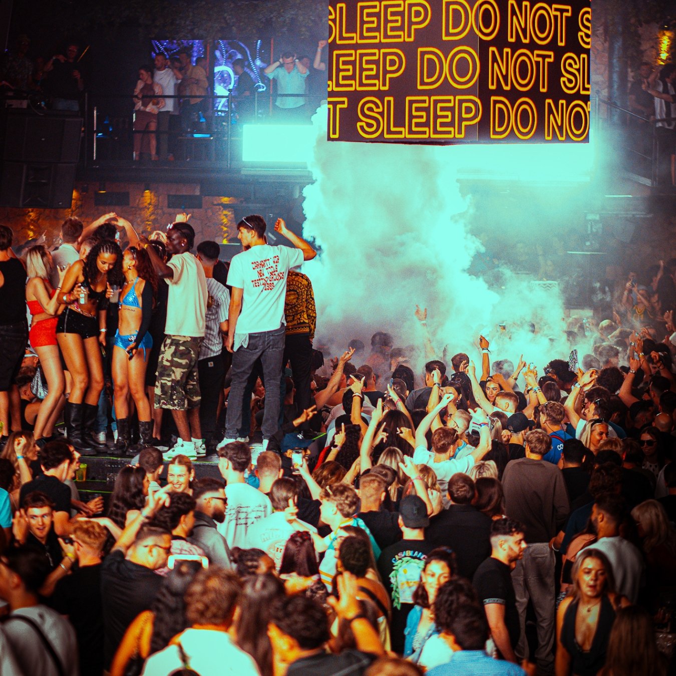 A crowded nightclub with people dancing, socializing, and enjoying the music. There are people on an elevated platform and a large electronic sign that reads 'SLEEP DO NOT SLEEP,' with colorful lights and smoke effects creating a lively atmosphere.