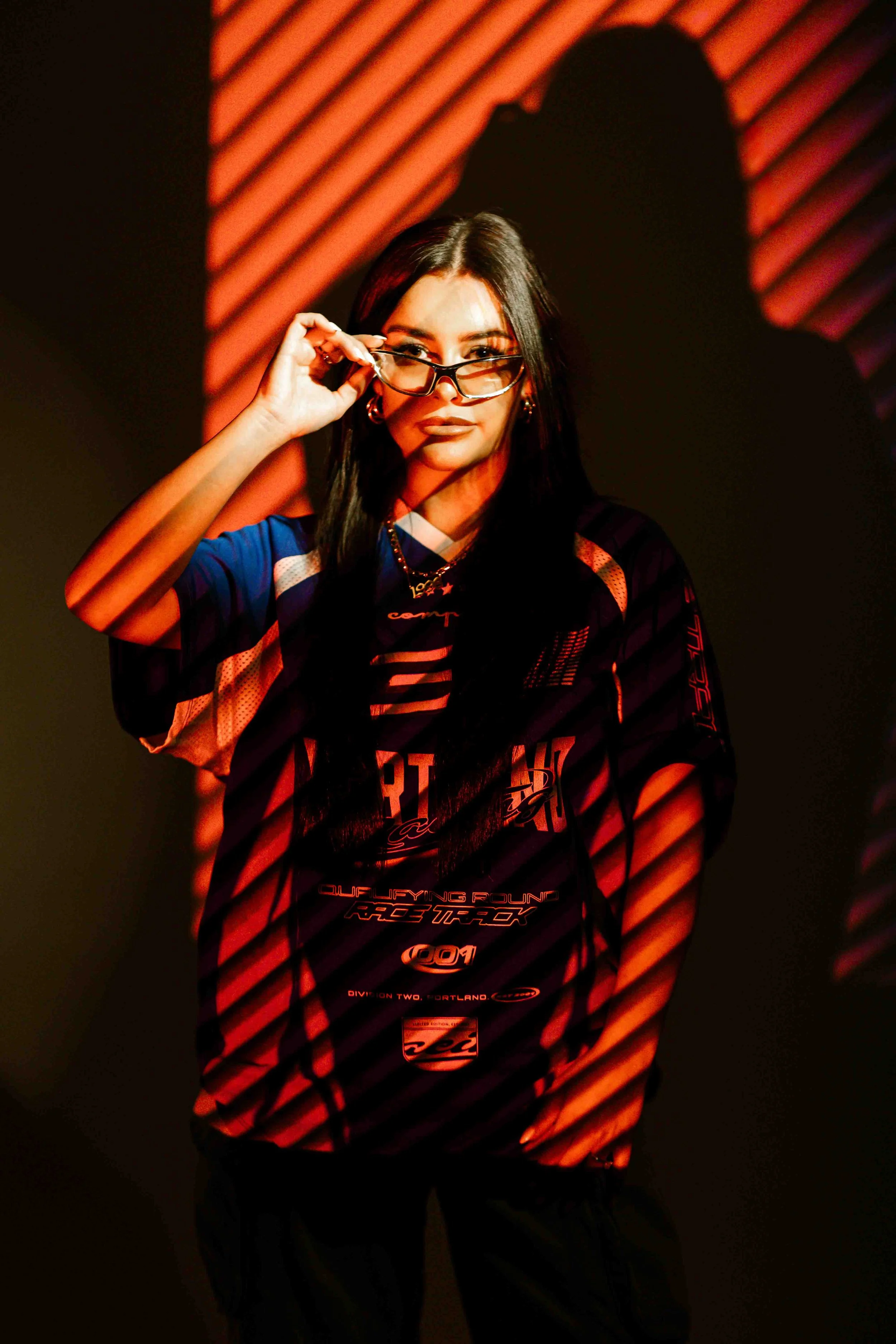 A woman with long dark hair and glasses, wearing a racing jersey, touches her glasses and looks at the camera under red and black diagonal lighting.