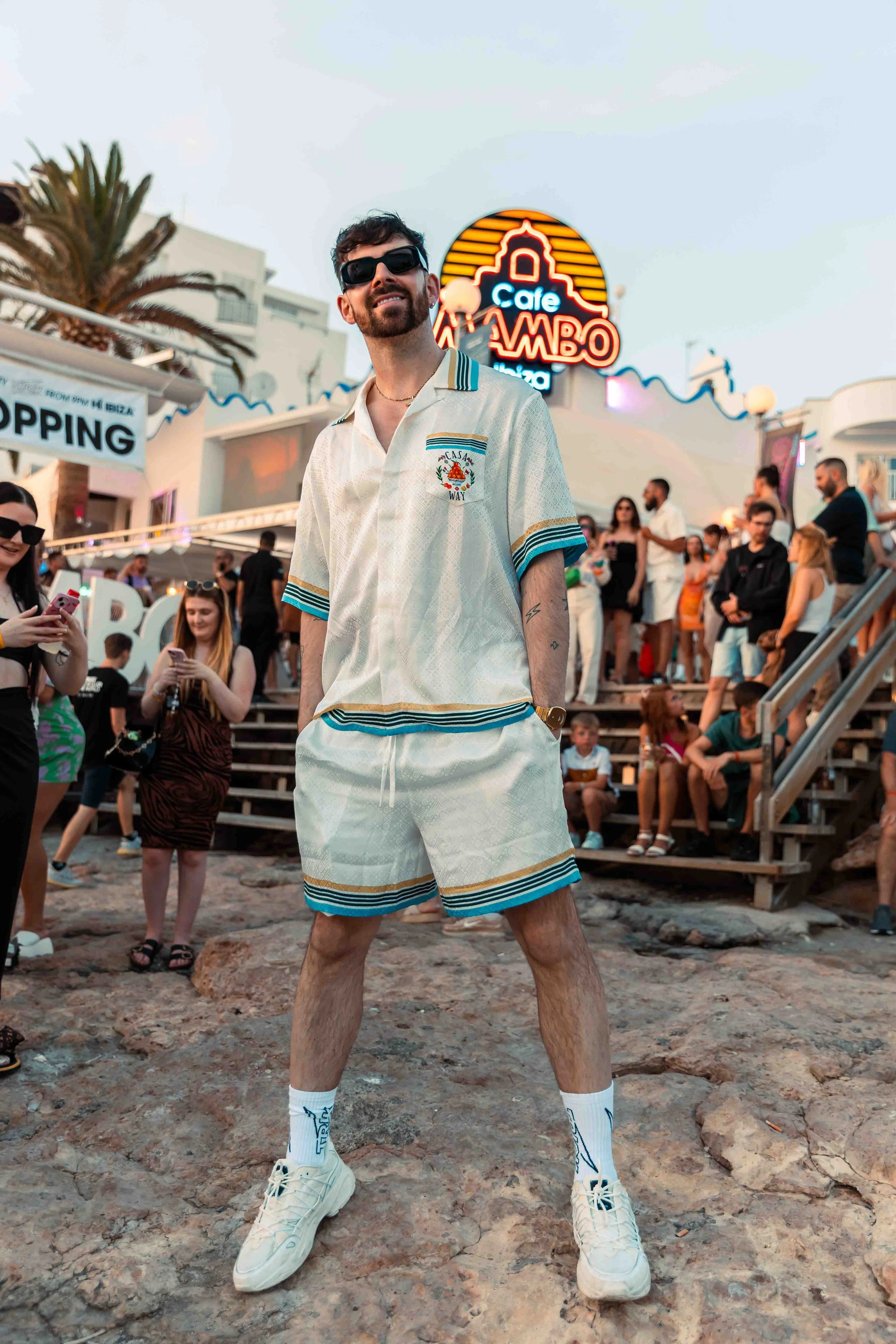 A young man with dark hair, a beard, and sunglasses standing on a rocky surface at a busy outdoor location with a crowd in the background. He is wearing a cream-colored matching shorts and shirt set with blue and yellow stripes, white sneakers, and w