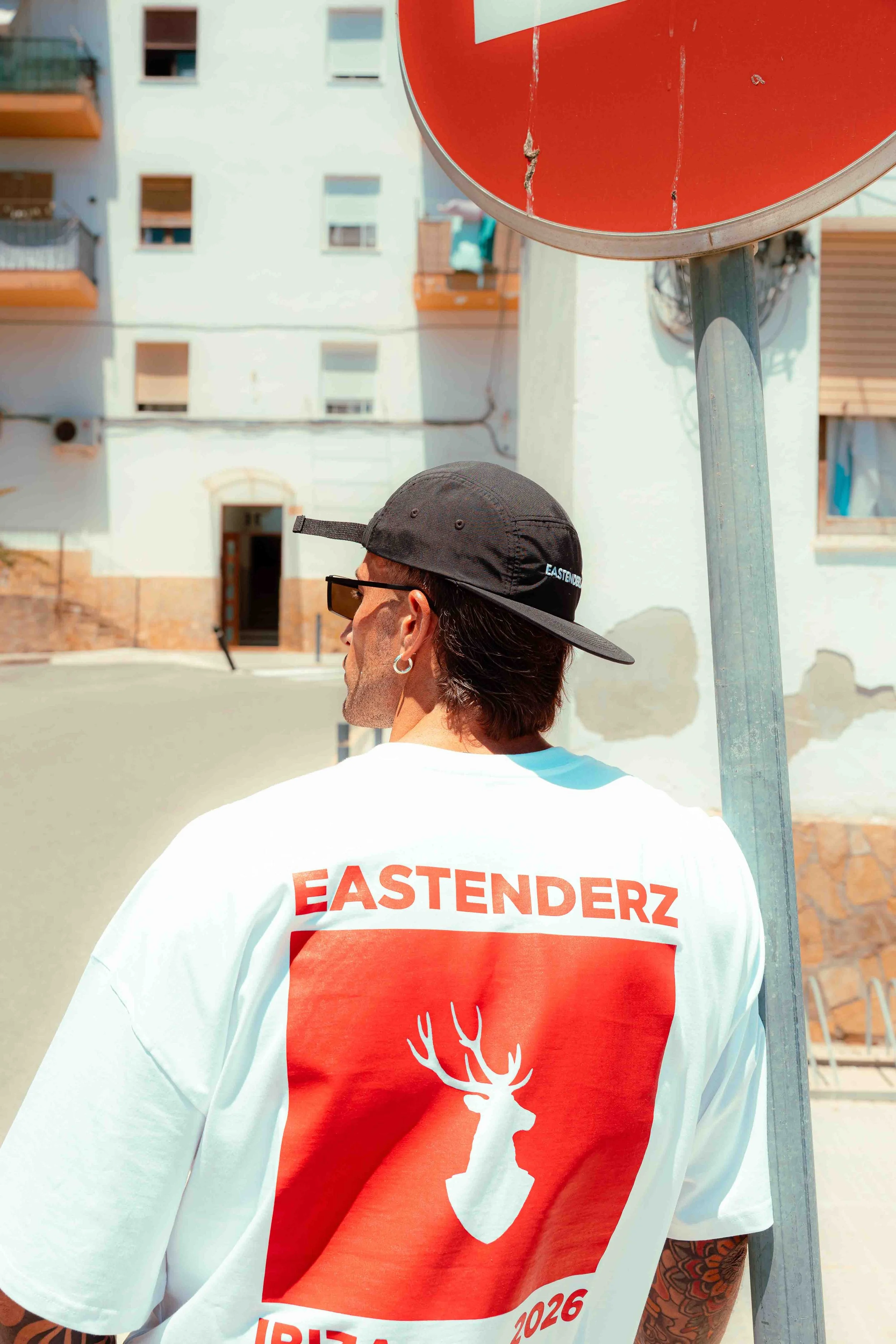 A man wearing a black cap, sunglasses, and a white T-shirt with a red graphic and the word 'EASTENDERZ' on the back. He is standing outdoors near a red circular sign and a building with balconies and windows.