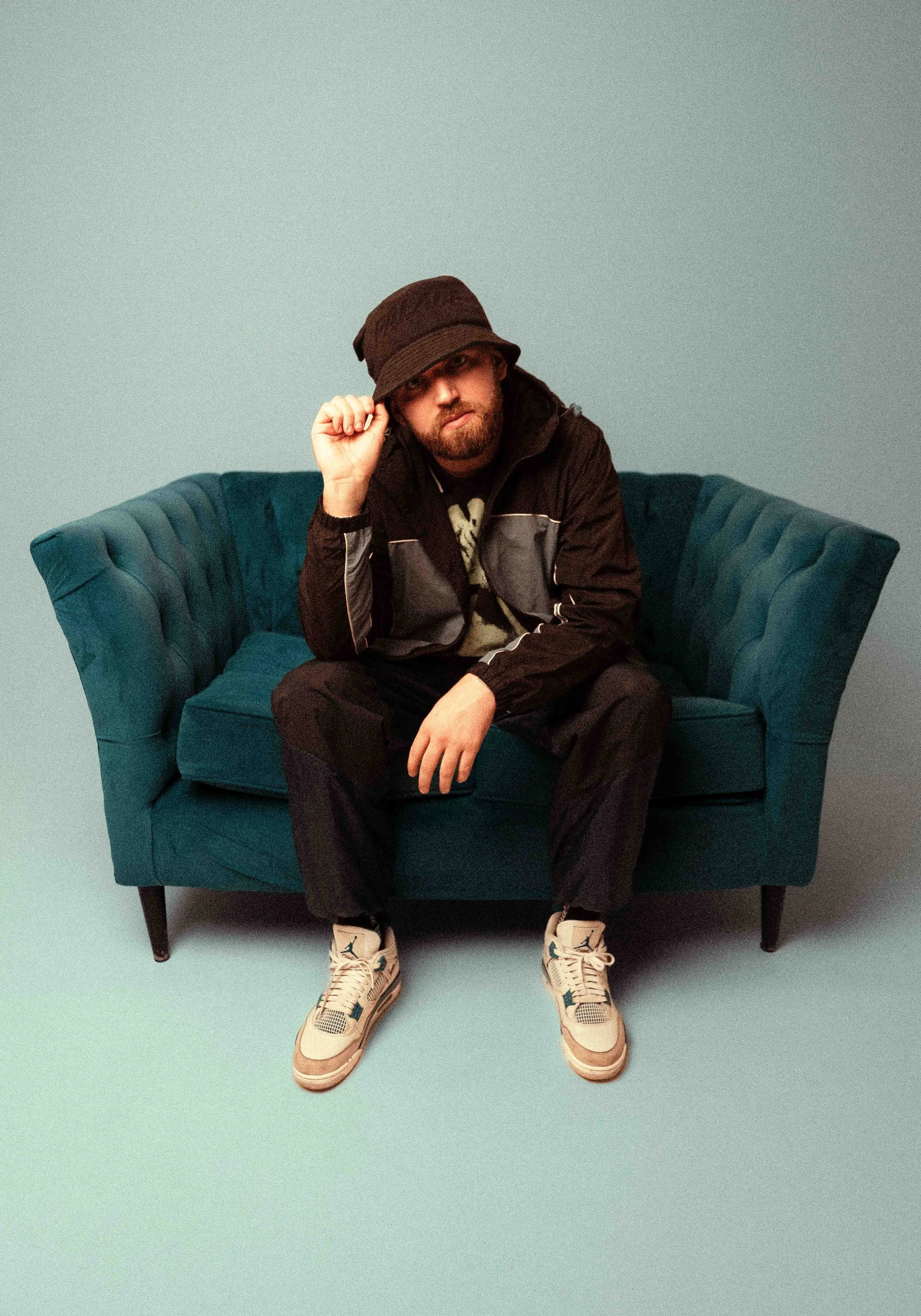 A man with a beard wearing a brown bucket hat, black jacket, and sneakers sitting on a teal velvet couch against a plain light blue-green background.