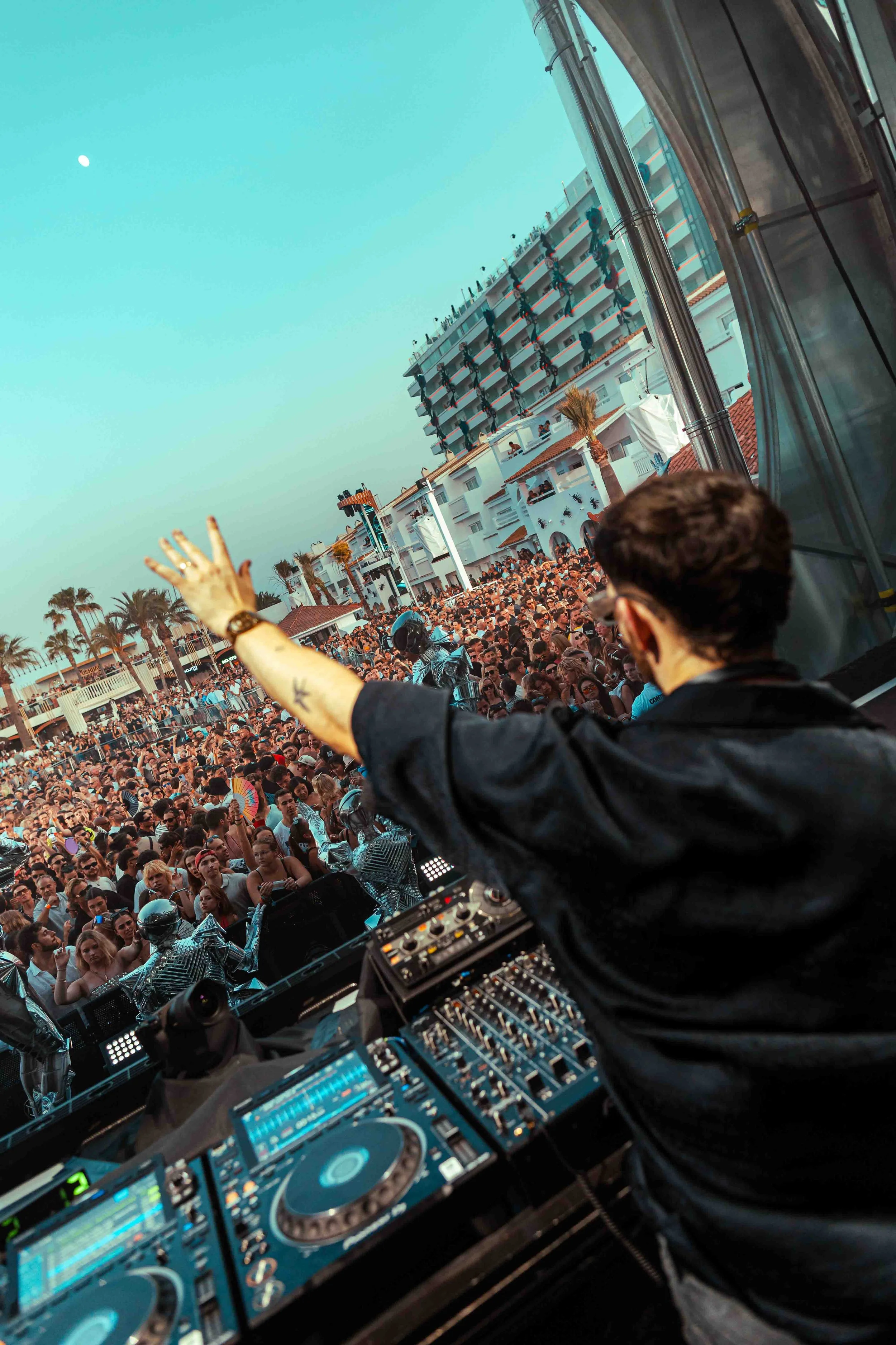 A DJ performing at an outdoor event, facing a large crowd with palm trees and a multi-story building in the background. The DJ's equipment is visible in the foreground.