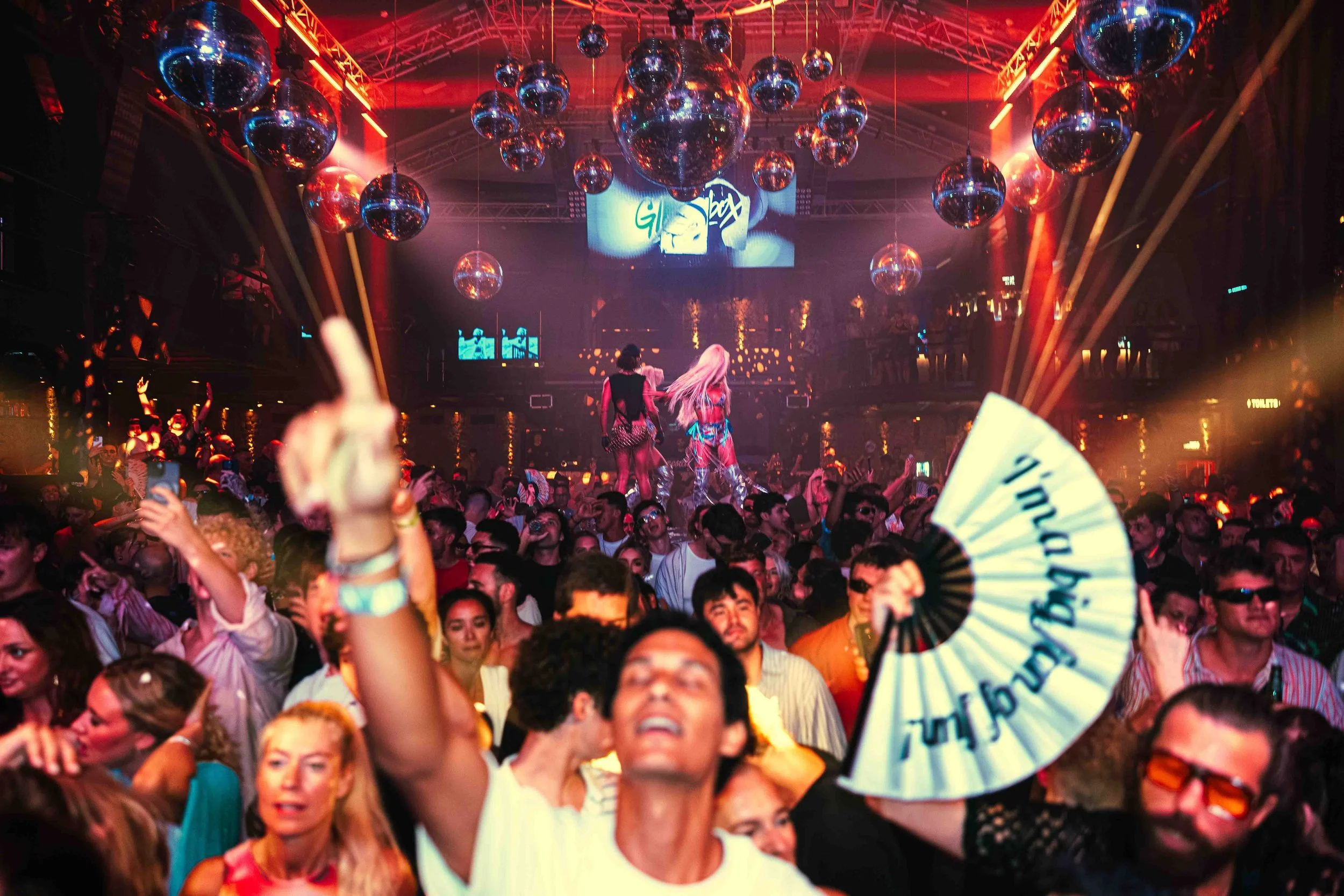 A crowded dance club or concert venue with multiple disco balls hanging from the ceiling and vibrant red and purple lighting. People are dancing and holding fans, with one person in the foreground raising their hand and a stage in the background with