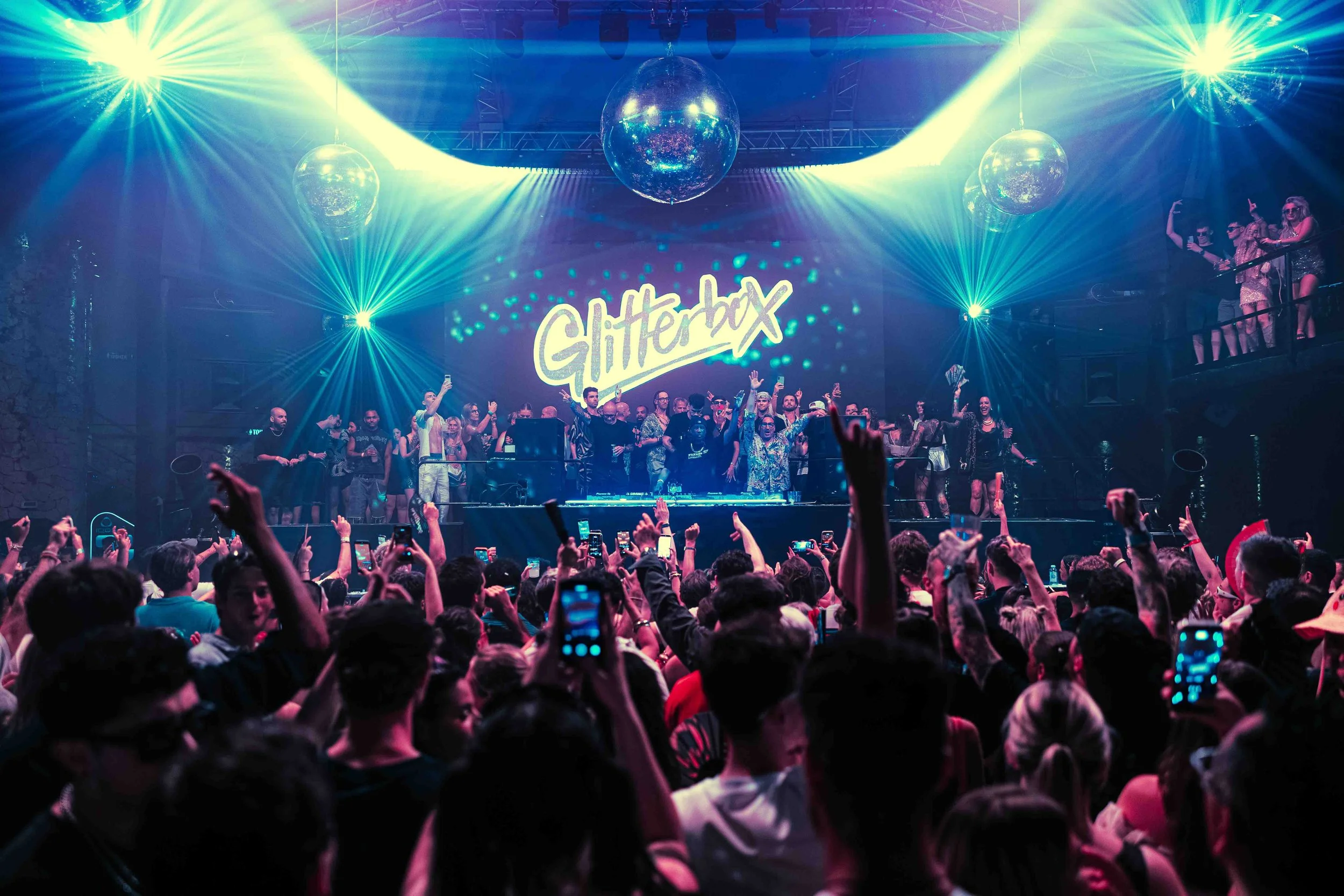 People dancing and enjoying themselves at a nightclub with colorful lights, a large screen displaying 'Glitterbox,' and multiple disco balls hanging from the ceiling.