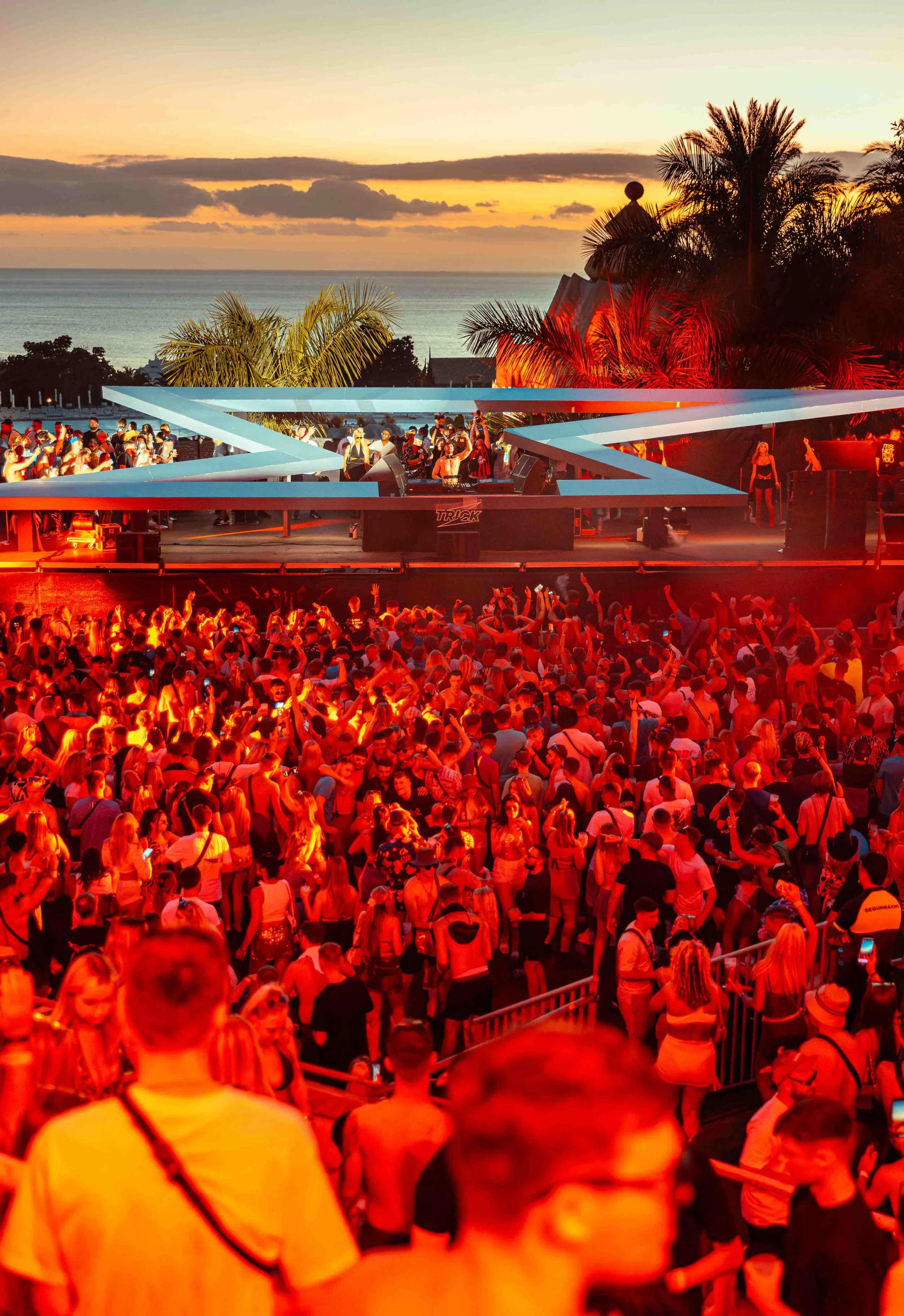 A large outdoor party or music festival at sunset with many people dancing and socializing. The stage is illuminated with red lights and surrounded by tropical palm trees. The background shows a sunset over the ocean with a cloudy sky.