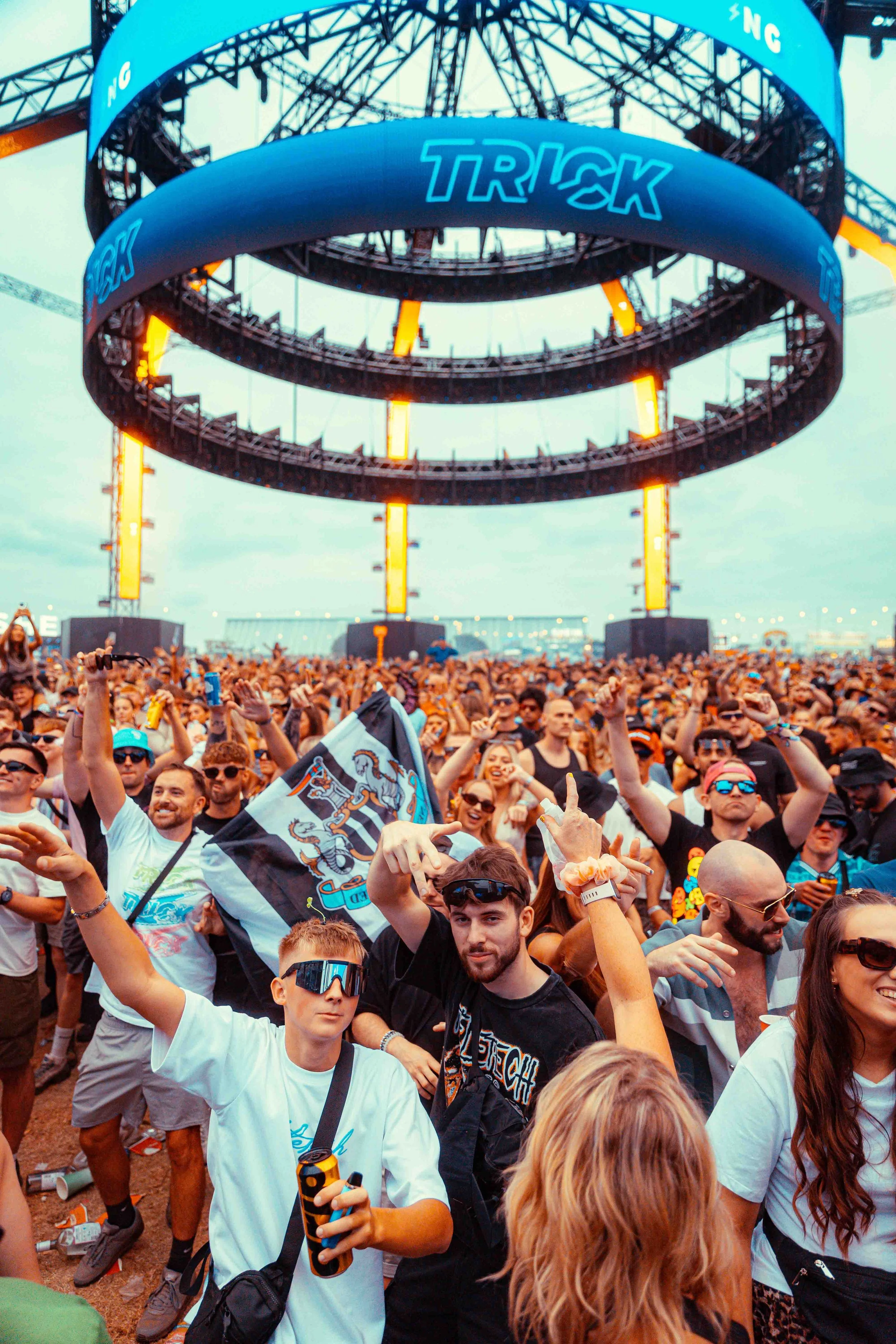 Crowd of people at an outdoor concert or festival. A large circular stage with a sign reading 'TRICK' is visible above the audience. Many attendees are raising their hands, some are holding drinks or flags, and some are wearing sunglasses. The sky is