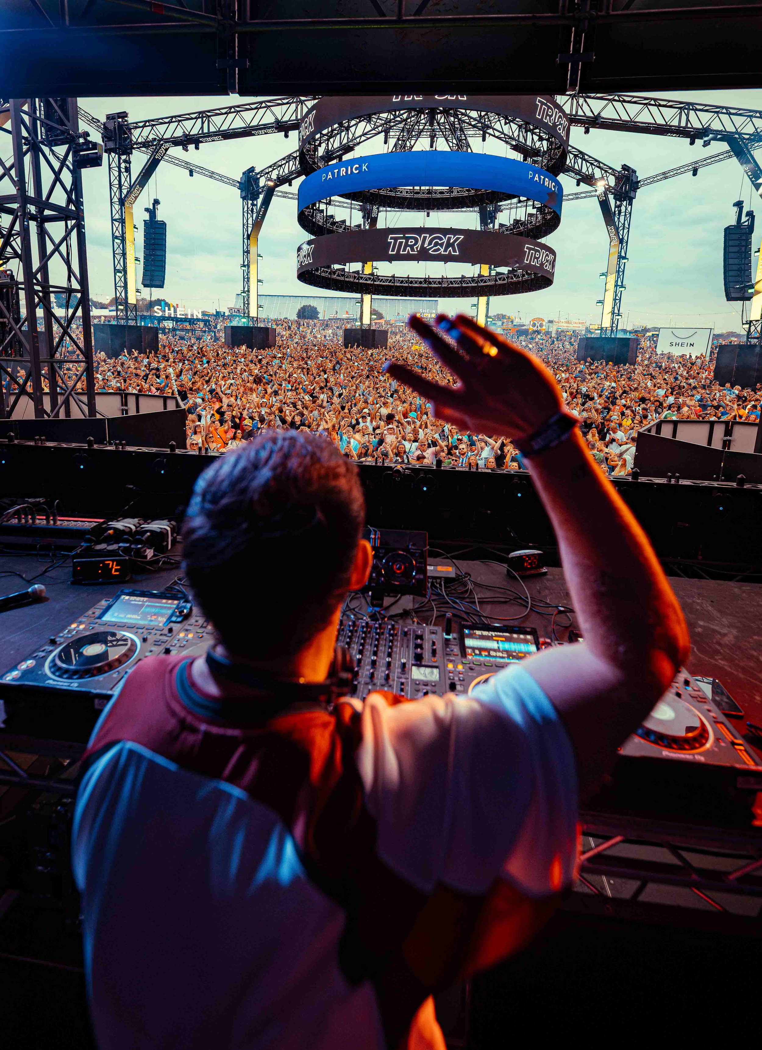 DJ performing on stage at a large outdoor music festival with a crowd of people dancing in front of the stage. The stage has a circular LED screen and metal trusses. The DJ is wearing headphones and a baseball cap.