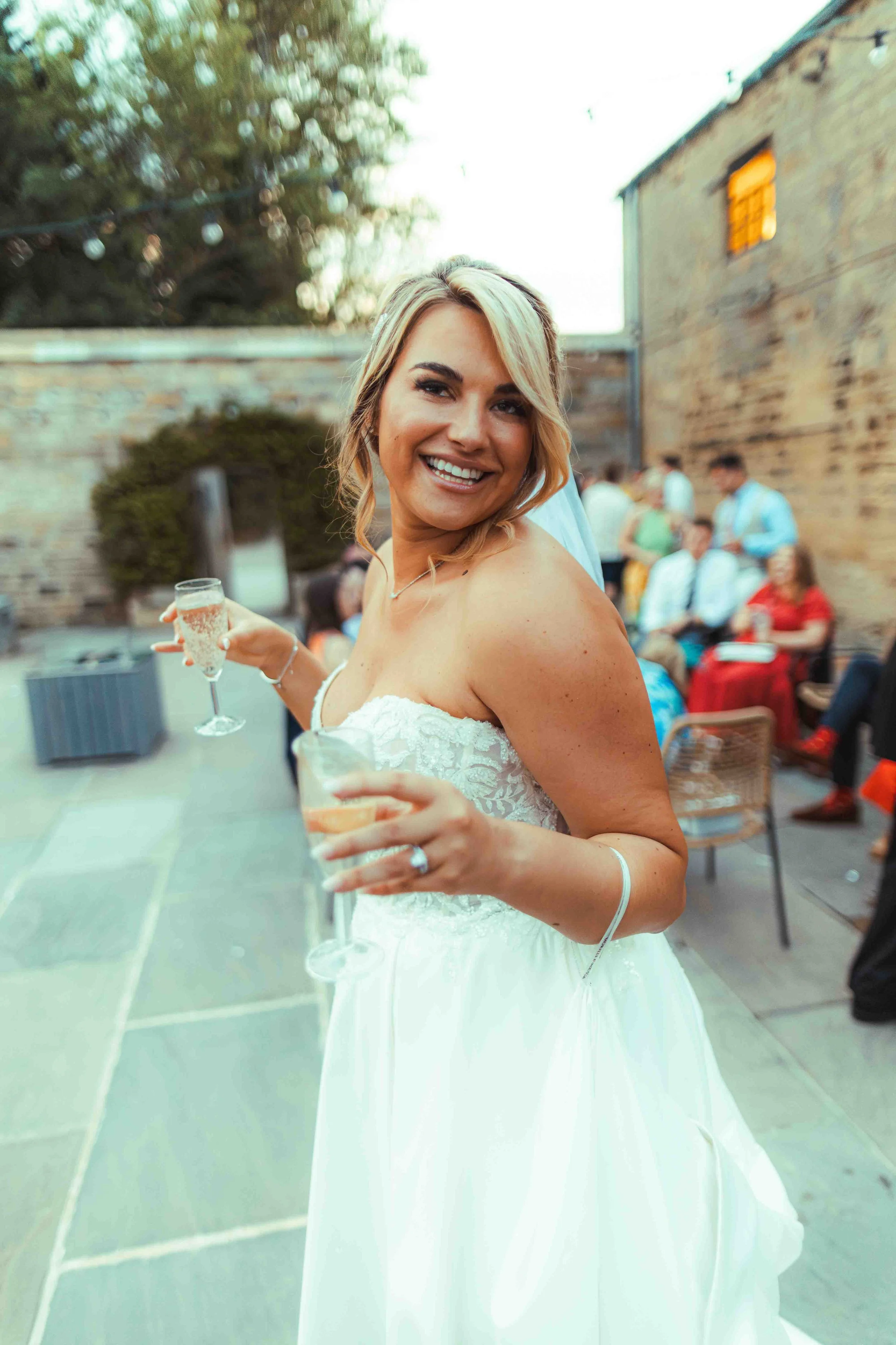 A smiling woman in a white dress holding a champagne glass at an outdoor gathering, with blurred guests seated in the background near a brick wall.