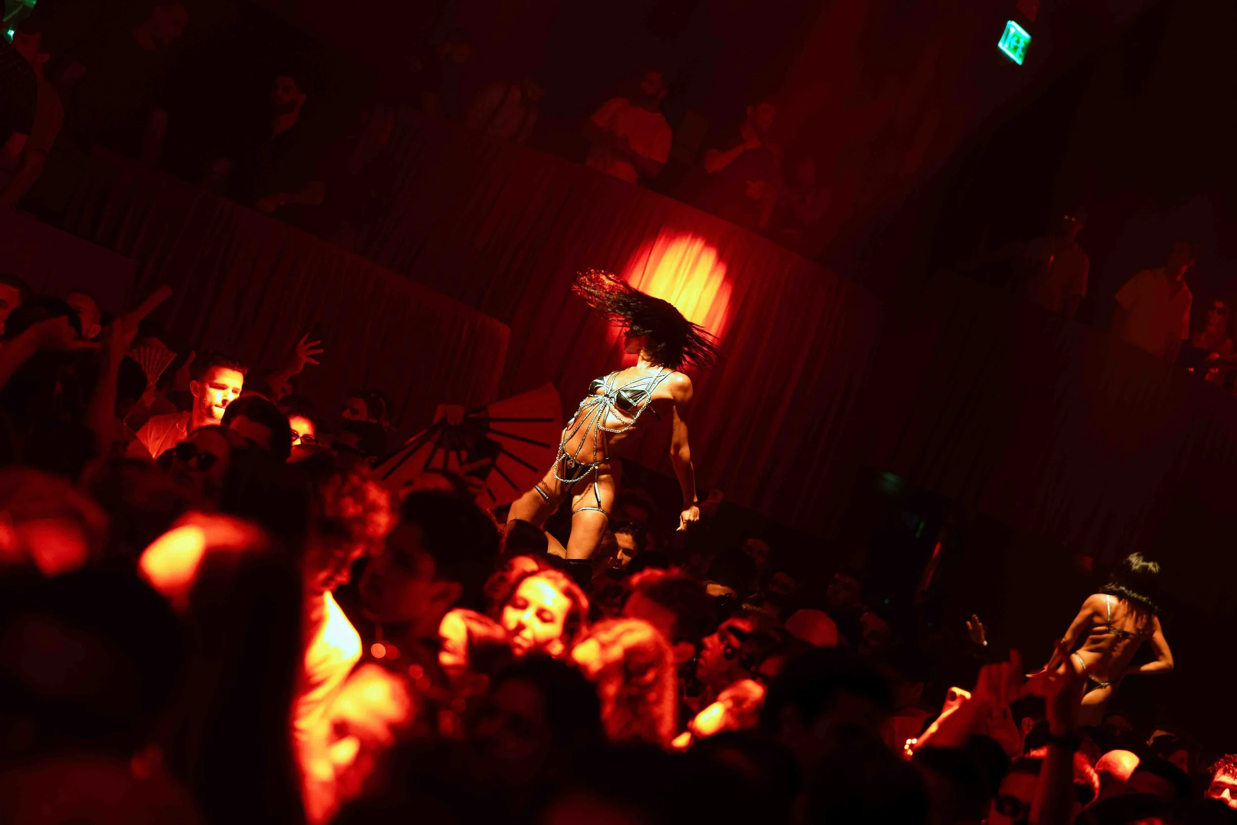 A performer dance on stage in front of a large crowd, with red lighting and an illuminated exit sign visible in the background.