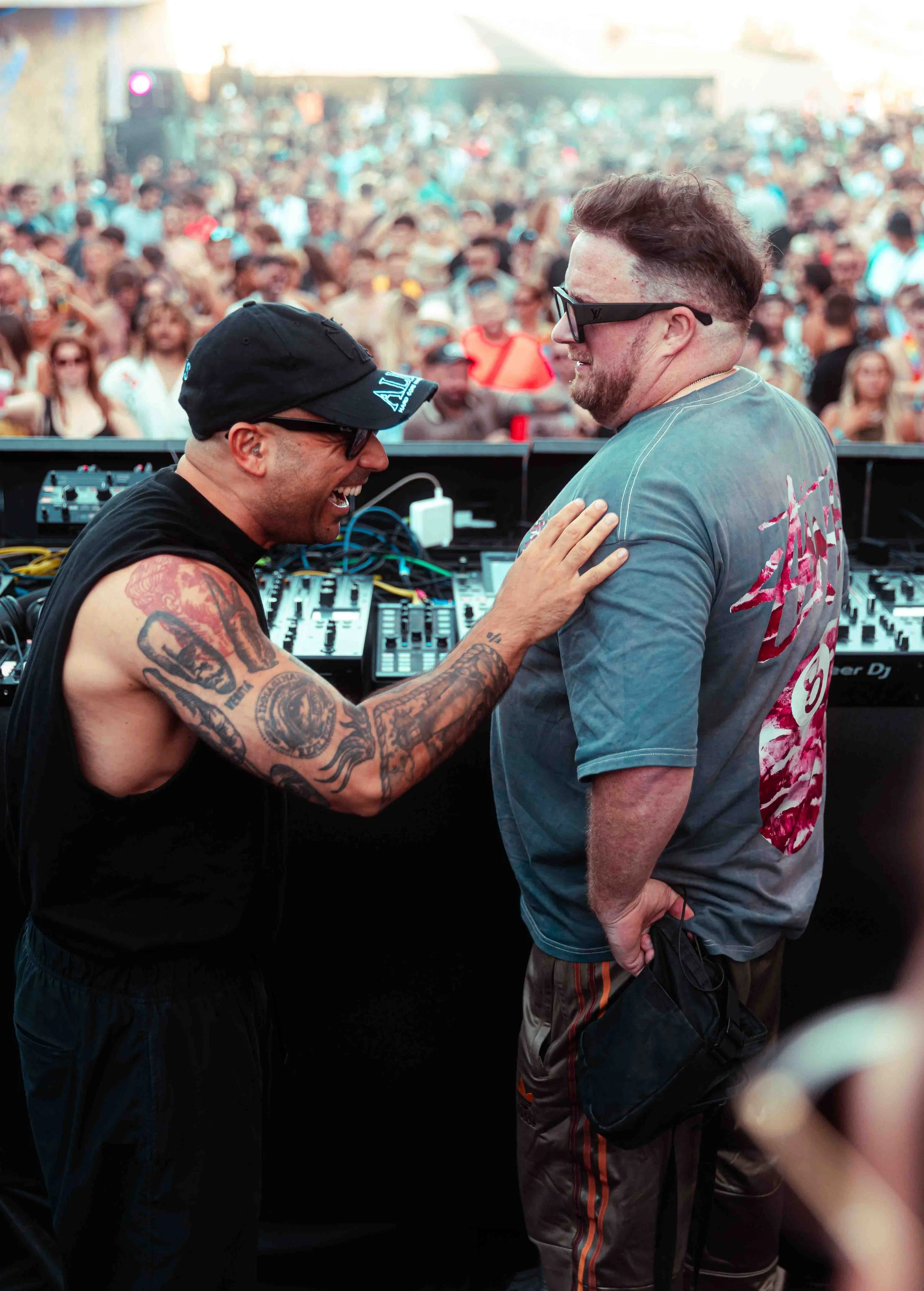 Two male DJs, one shirtless with tattoos and wearing a black cap, and the other in a gray T-shirt and sunglasses, smiling and interacting at an outdoor music event with a large crowd in the background.