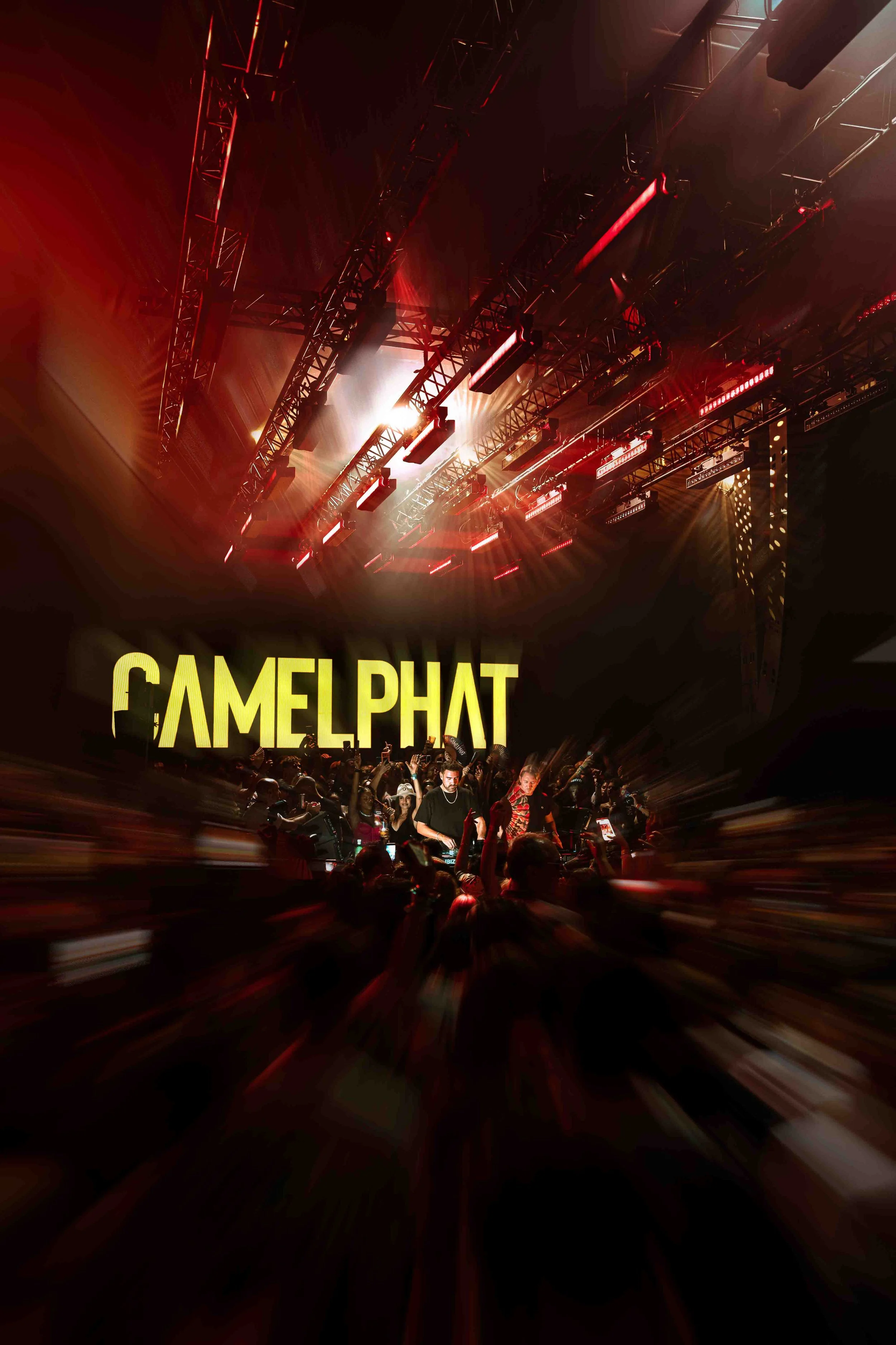 Crowd at a music event with a stage and large illuminated sign reading 'GAMELPHAT' in the background, stage lighting with red and white lights and trusses overhead.