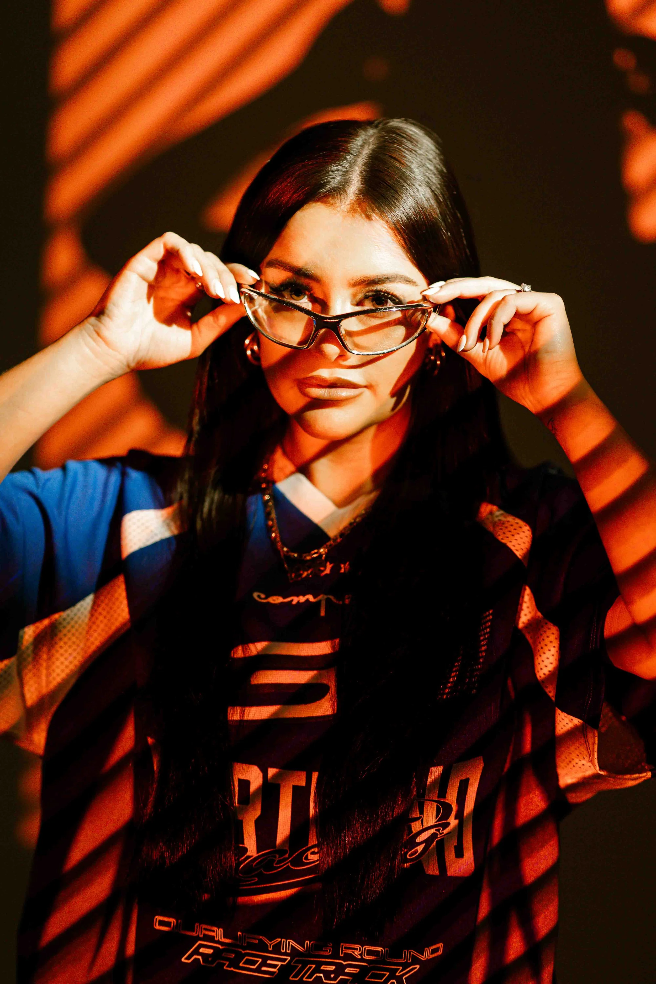 Eliya Jaia with dark hair and glasses looks over her sunglasses, with orange and black lighting highlighting her face, wearing a sports jersey.