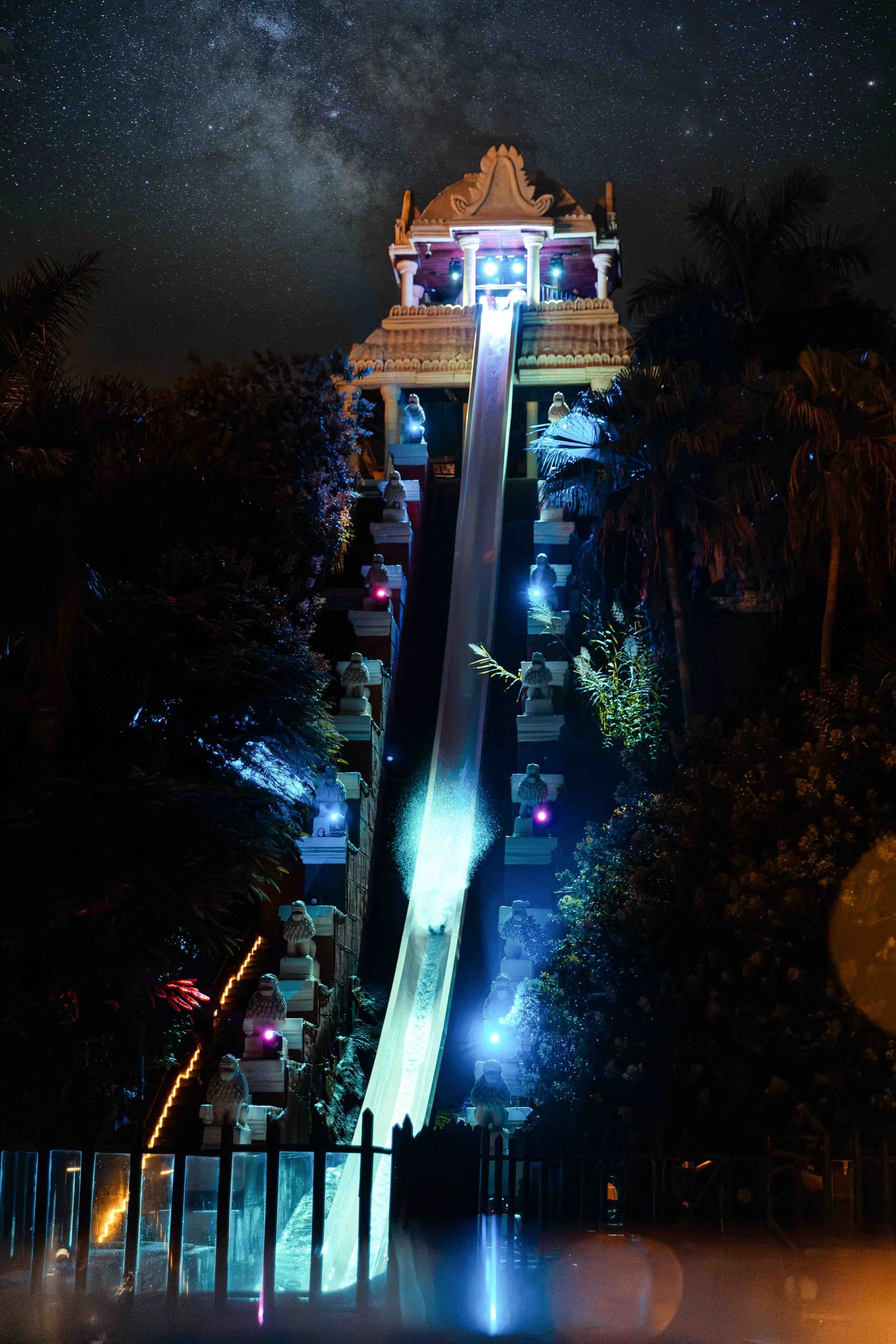 Nighttime view of a large amusement park water slide with a steep drop, illuminated by colorful lights, with trees and stars visible in the sky.