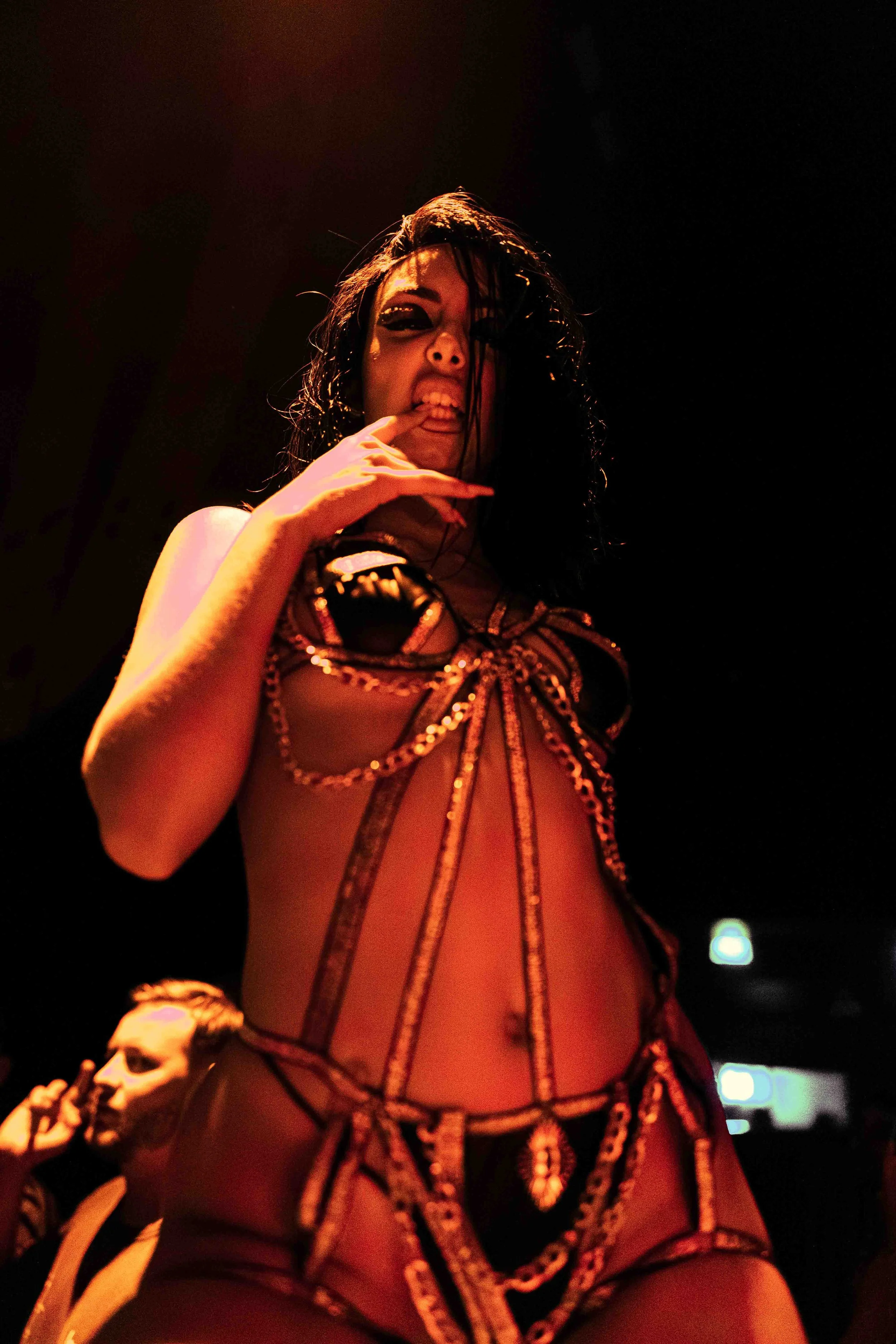 A woman with wet black hair making a playful face, sticking out her tongue, and touching her lips with her finger, wearing a black and gold revealing outfit with chain detailing, in a dark setting.