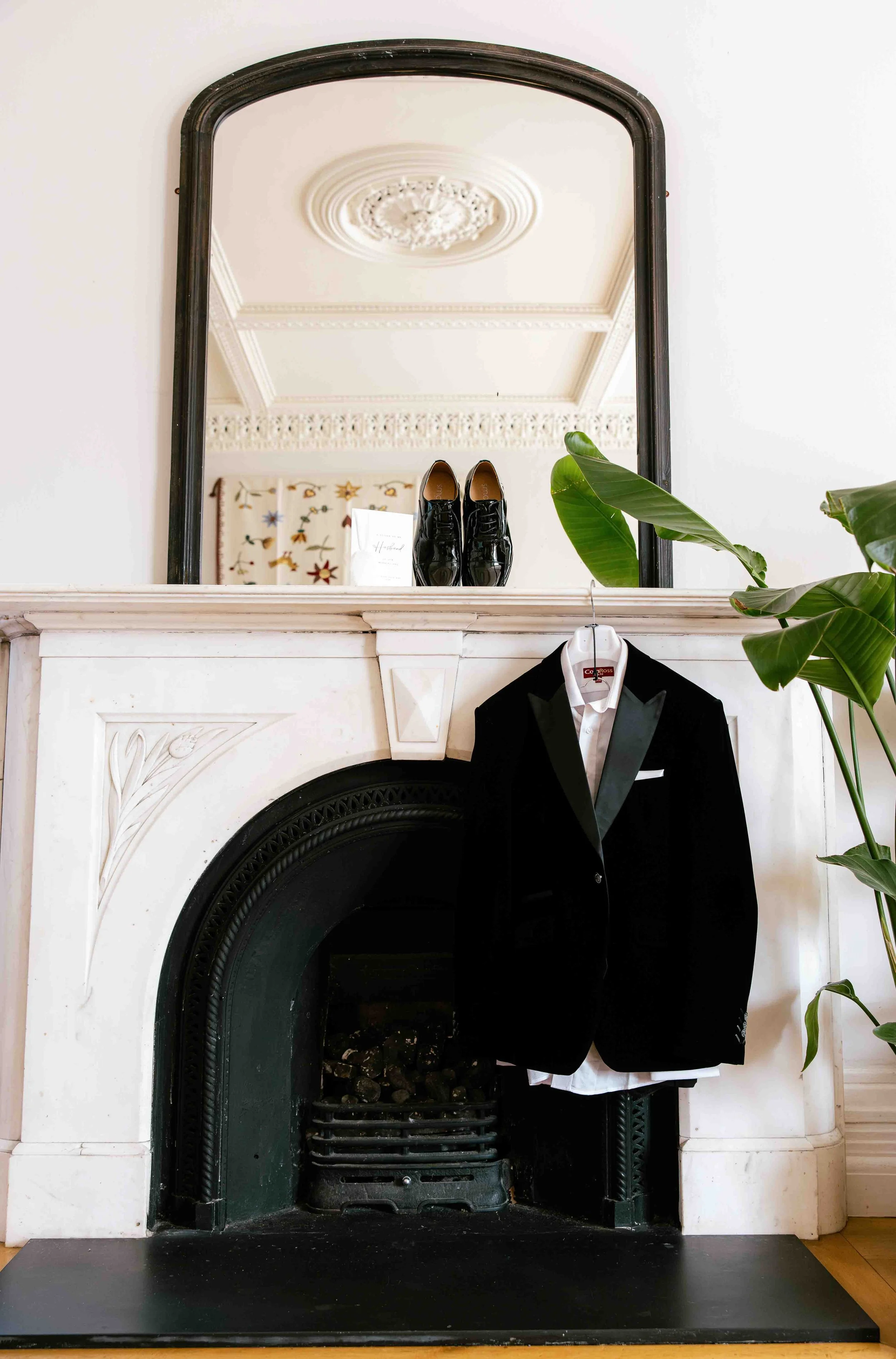 A black tuxedo hanging on a fireplace mantel with a white shirt underneath, a pair of shiny black shoes on the mirror shelf, a large mirror above the mantel reflecting the ceiling, a leafy green plant to the right, and a decorative pillow with bird and floral embroidery in the background.