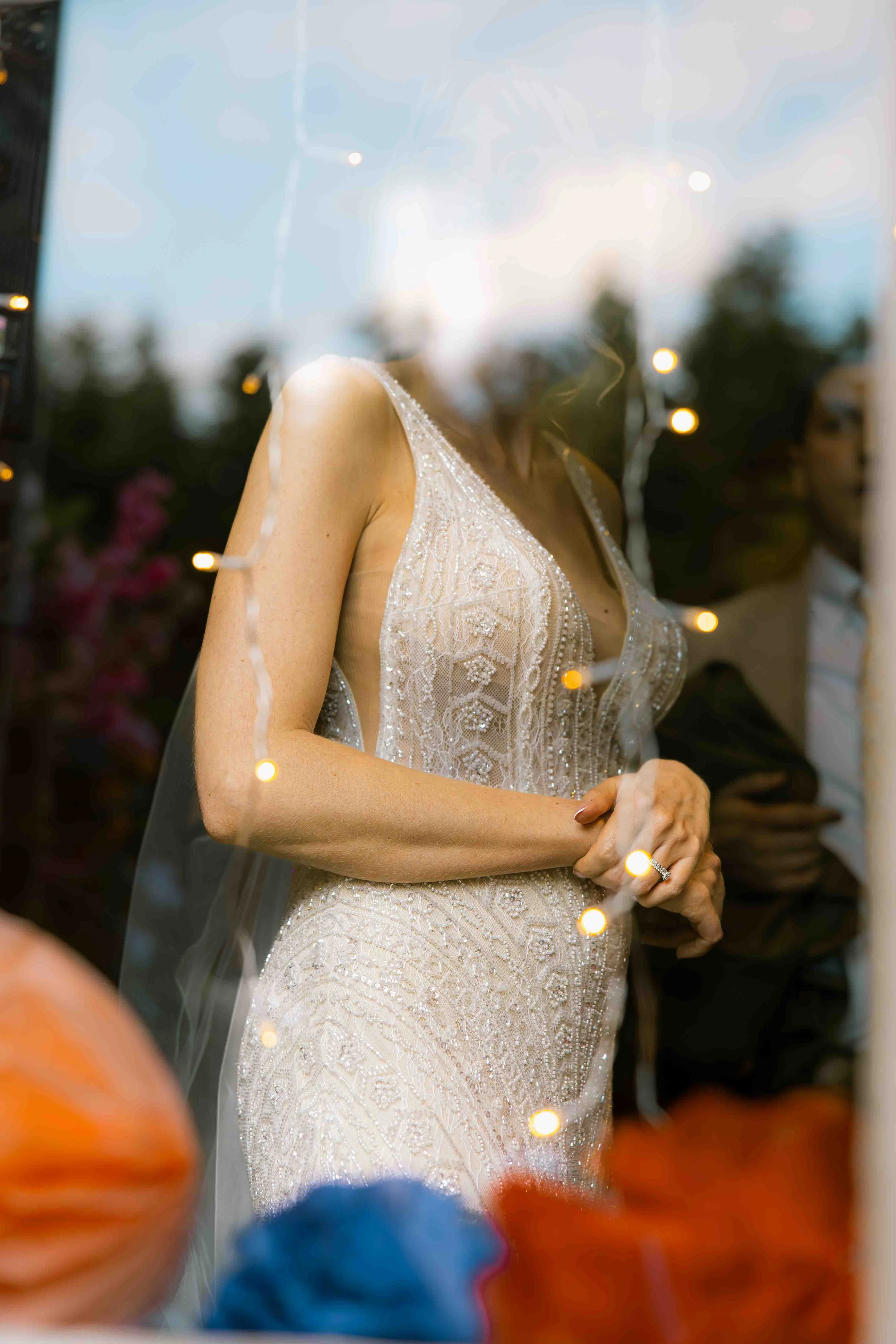 Close-up of a woman in a wedding dress with intricate beading and lace, standing outdoors at sunset or evening, with blurred trees and string lights in the background.