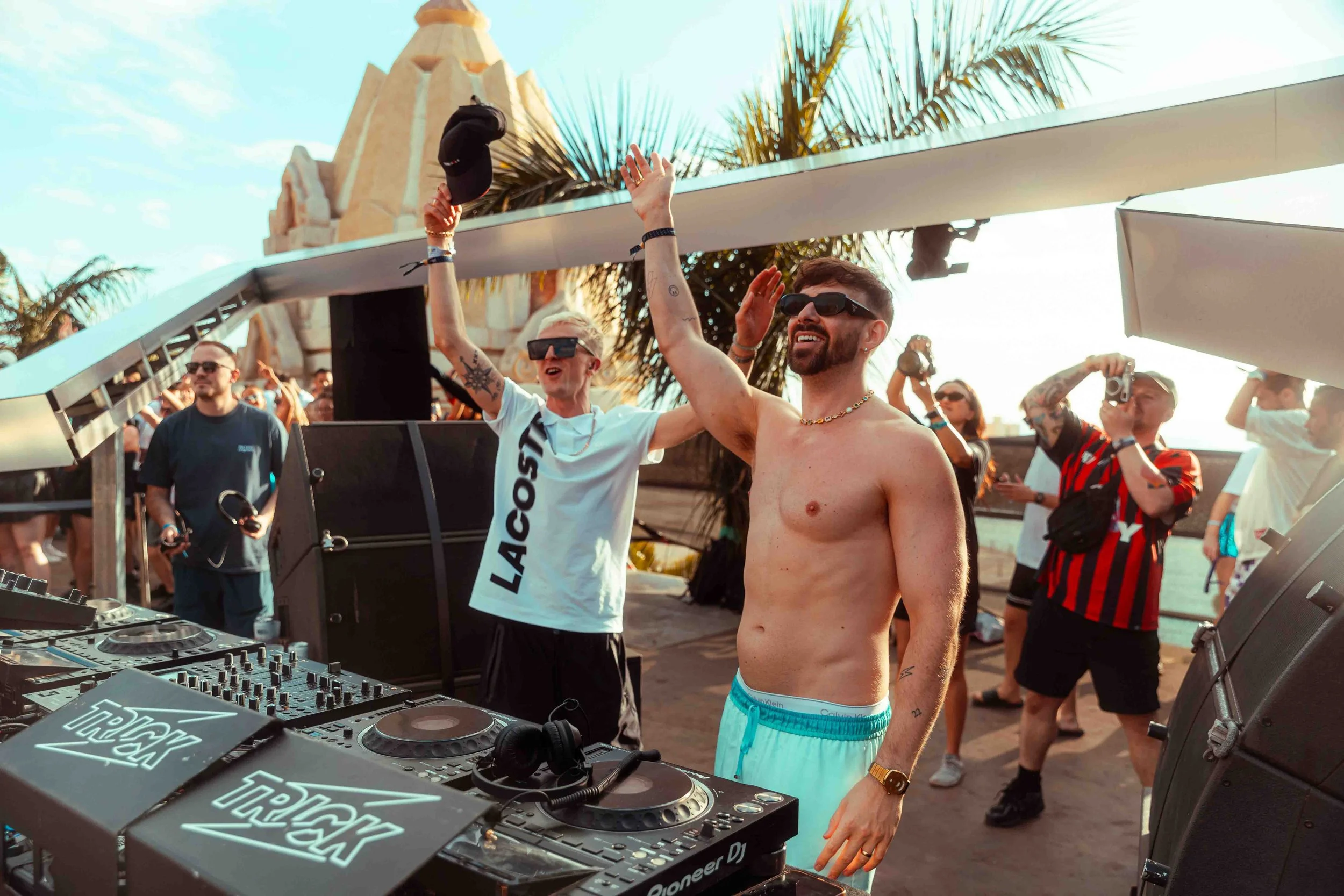 People dancing and enjoying music at an outdoor event with a DJ booth in the foreground and a palm tree in the background.