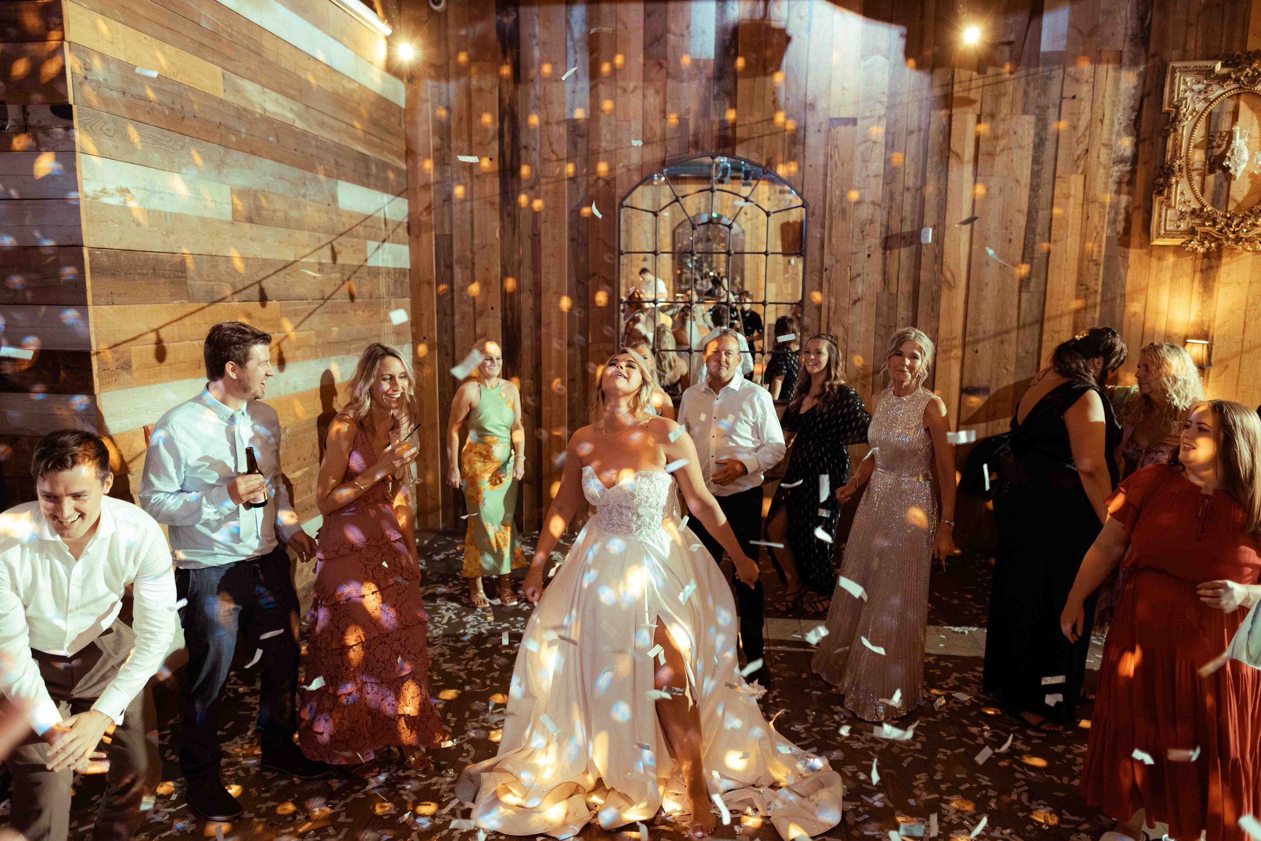 People celebrating at a wedding reception with confetti falling, dancing, and smiling in a rustic indoor venue with wooden walls and decorative mirrors.
