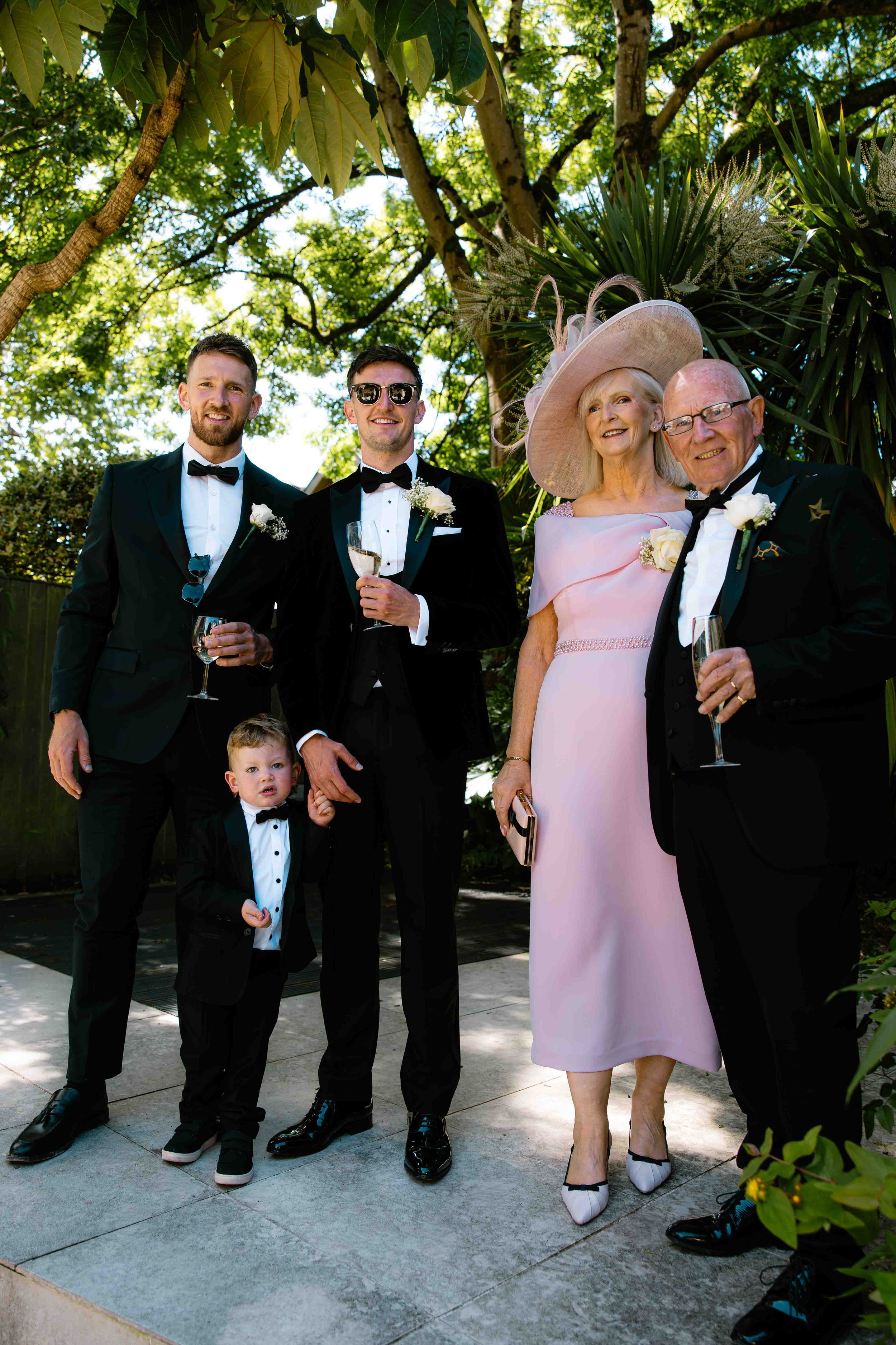 A group of five people, three men, one woman, and one child, dressed in formal wedding attire, standing outdoors under green tree foliage and sunlight, smiling and holding champagne glasses.
